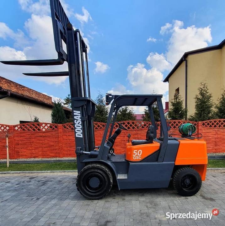 Wózek widłowy marki Doosan, model 50 GSC, pomarańczowo-szary, zaparkowany na kostce brukowej przed budynkiem mieszkalnym i drewnianym ogrodzeniem.