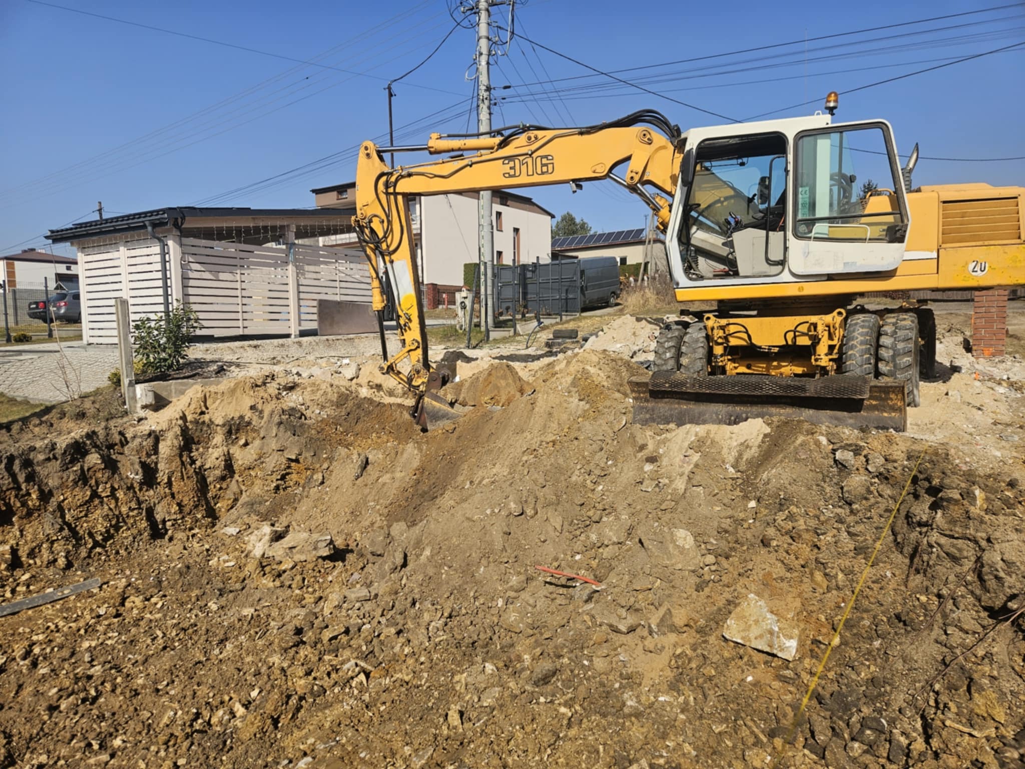 Żółto-biała koparka Liebherr 316 przy wykopie w ziemi, widoczne budynki w tle i błękitne niebo.