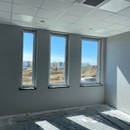 SV-BUD Serhii Vovk - Wnętrze pomalowanego na szaro pomieszczenia z trzema oknami. Podłoga zabezpieczona folią malarską. Widoczne gniazdka elektryczne i kratka wentylacyjna.