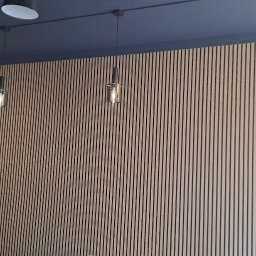 SV-BUD Serhii Vovk - Ściana wyłożona pionowymi panelami z drewna, podwieszane lampy. Ciemny sufit z widoczną instalacją wentylacyjną. Fragment osoby w lewym dolnym rogu.