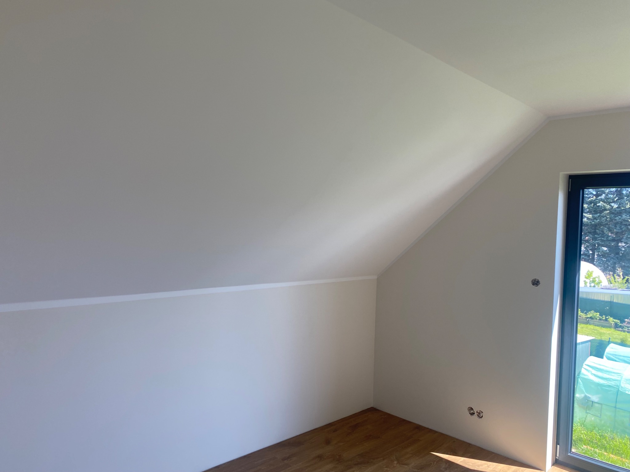 Poddasze po remoncie: białe ściany, drewniana podłoga, okno z widokiem na zieleń. Gładkie powierzchnie, brak mebli, widoczne gniazdka elektryczne.