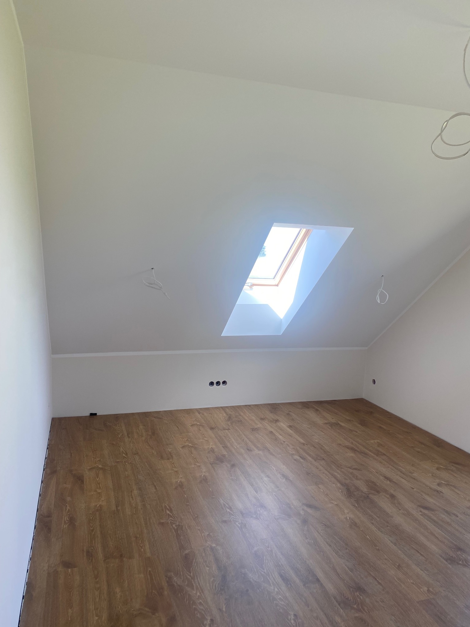Poddasze po remoncie: świeżo położona podłoga z paneli w odcieniach brązu, białe ściany i sufit z oknem dachowym wpuszczającym naturalne światło. Widoczne gniazdka elektryczne.