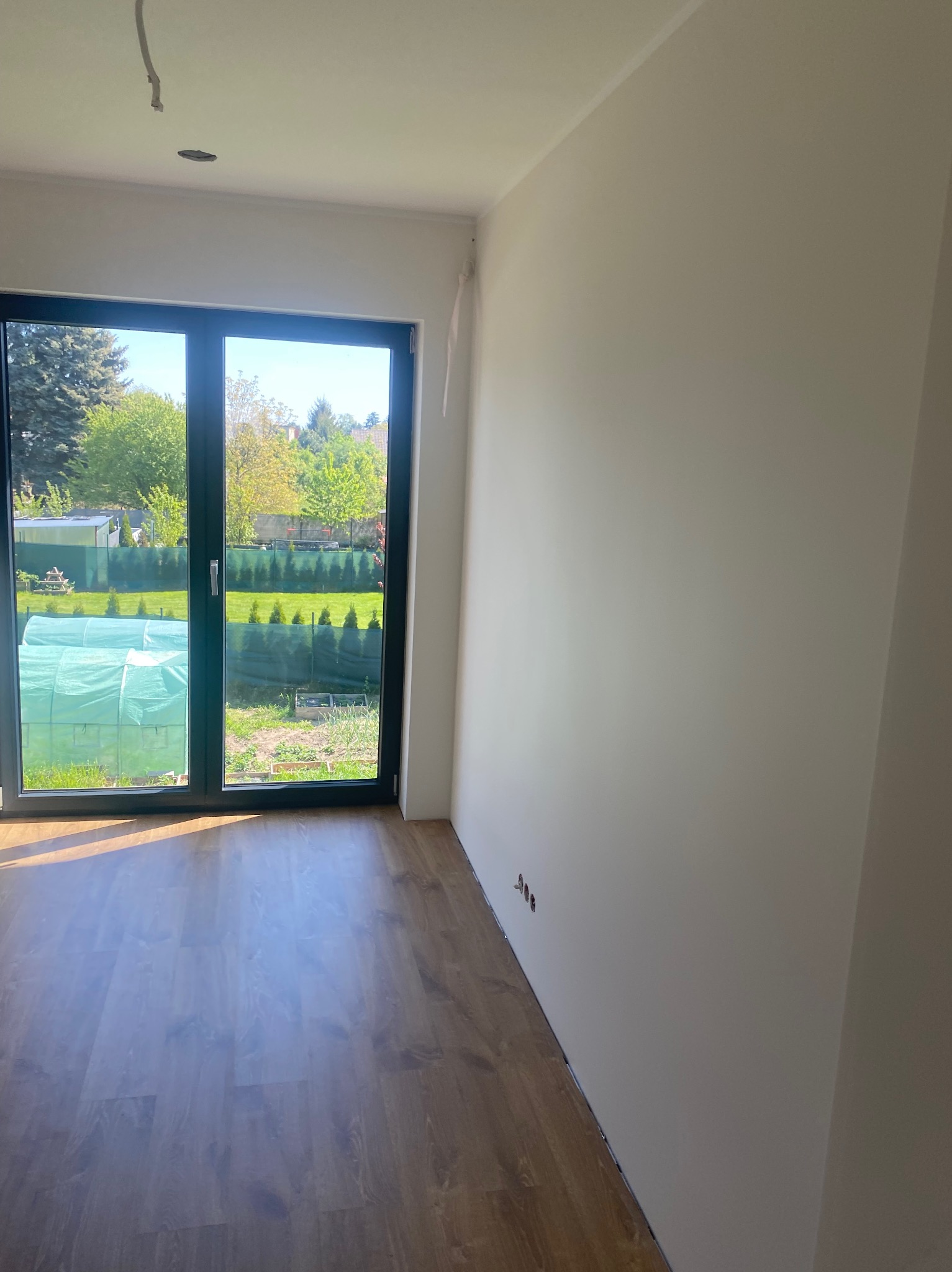 Pomalowane na biało wnętrze pokoju z panelami podłogowymi i dużym oknem balkonowym z widokiem na ogród. Widoczne gniazdka elektryczne i wyprowadzony kabel na suficie.