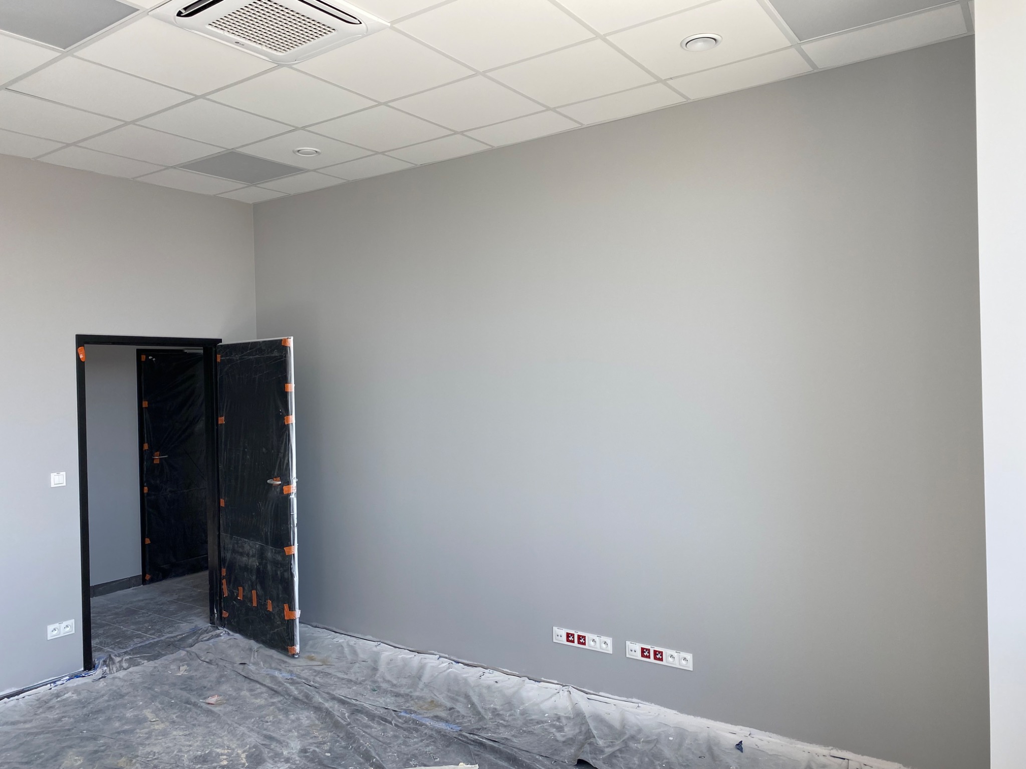 Szare ściany w pomieszczeniu biurowym po malowaniu, zabezpieczone drzwi, folia ochronna na podłodze, widoczne gniazdka elektryczne.