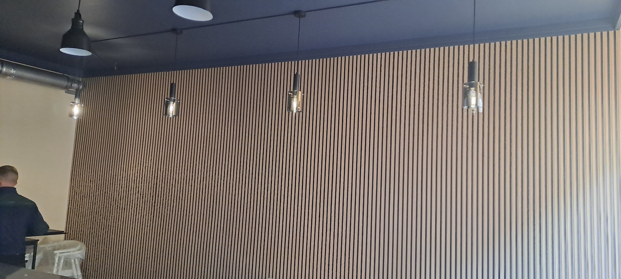 Ściana wyłożona pionowymi panelami z drewna, podwieszane lampy. Ciemny sufit z widoczną instalacją wentylacyjną. Fragment osoby w lewym dolnym rogu.
