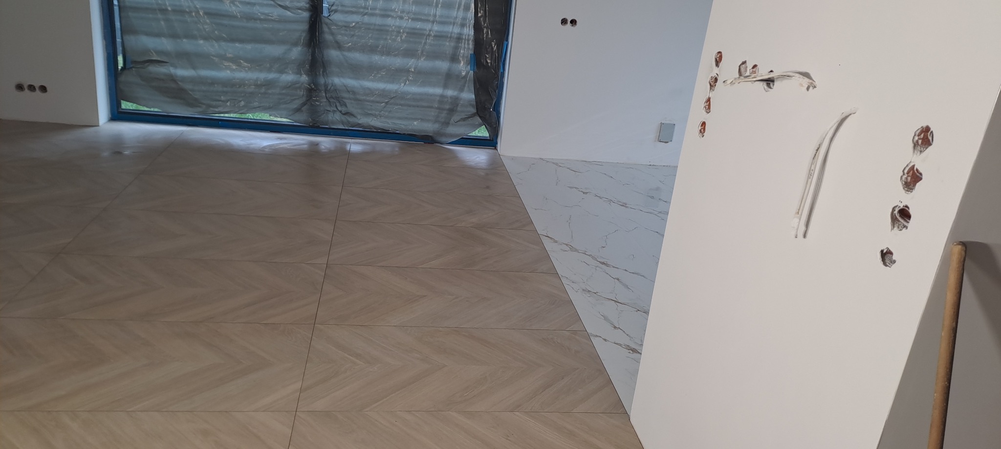 Ułożona podłoga z płytek w jodełkę i fragment ściany z instalacją elektryczną. Widoczne dwa rodzaje płytek podłogowych. Okno zabezpieczone folią.