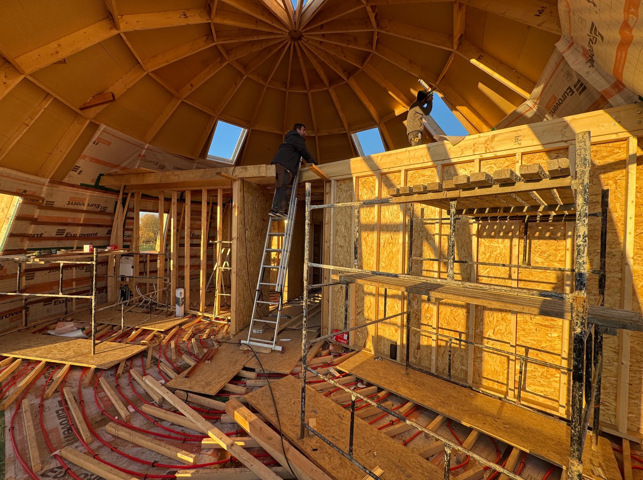 Wnętrze domu w trakcie budowy z drewnianą konstrukcją kopuły, instalacją ogrzewania podłogowego i pracownikami na rusztowaniu. Widoczne okna dachowe i elementy izolacji.