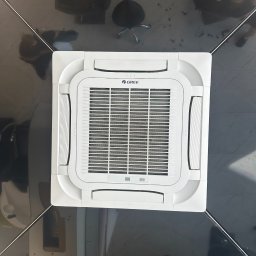 ECOTEMP SOLUTIONS SPÓŁKA Z OGRANICZONĄ ODPOWIEDZIALNOŚCIĄ - Sufitowa kaseta klimatyzacyjna marki Gree w pomieszczeniu biurowym z ciemną podłogą i meblami, widok z dołu.