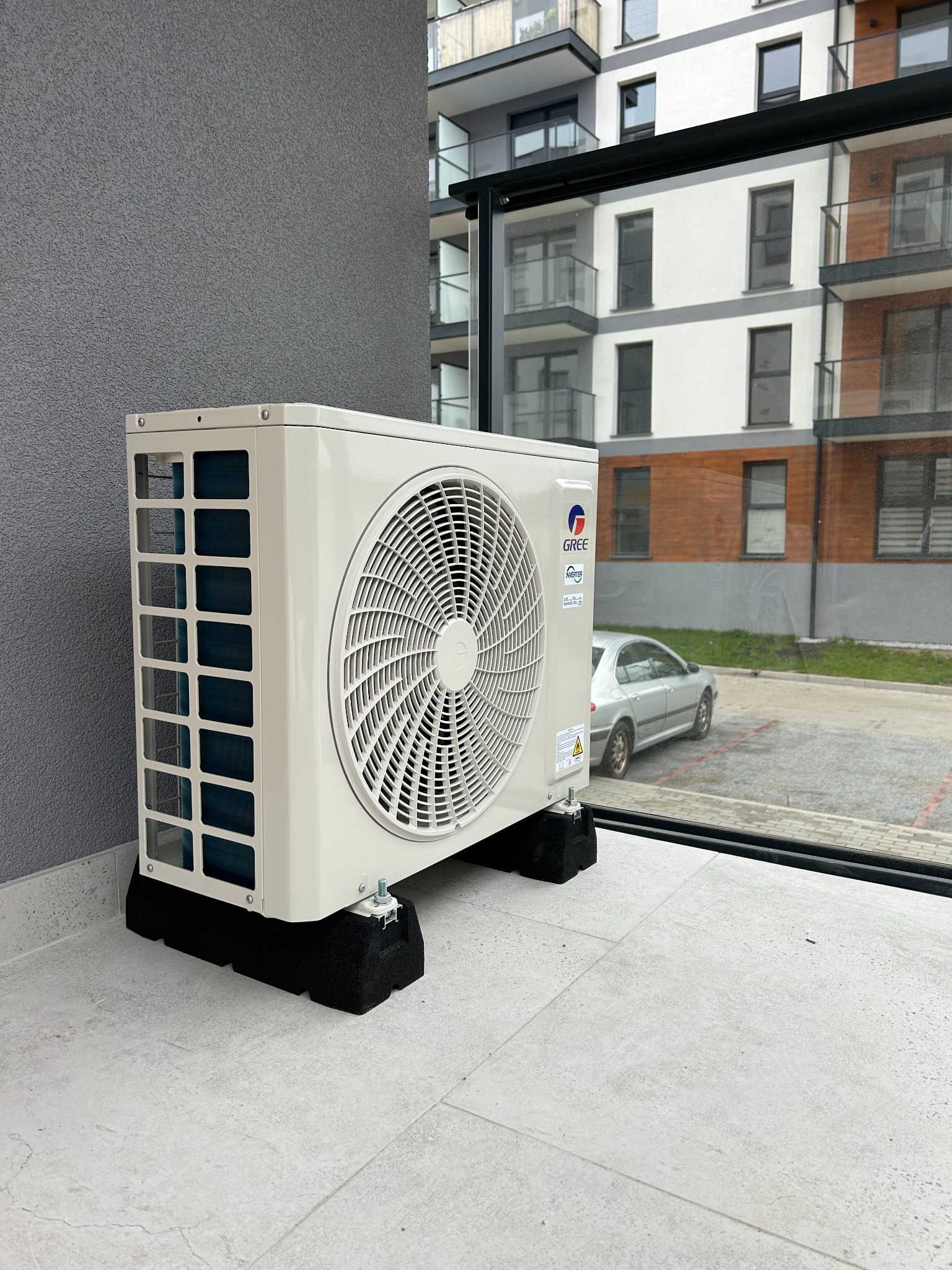 Zewnętrzna jednostka klimatyzacji marki Gree zamontowana na balkonie nowoczesnego apartamentowca, widoczna przez szybę balkonu, z szarym samochodem w tle.