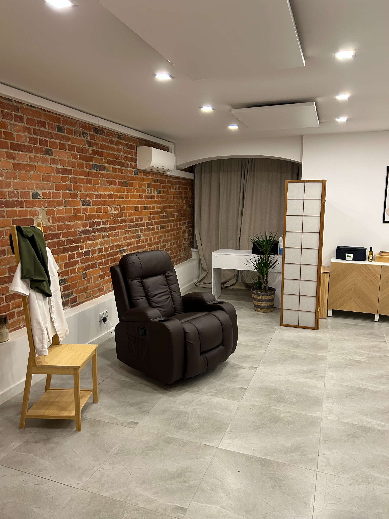 Wnętrze pokoju z odsłoniętą ceglaną ścianą, brązowym fotelem relaksacyjnym, i wiszącą jednostką klimatyzacyjną na ścianie.