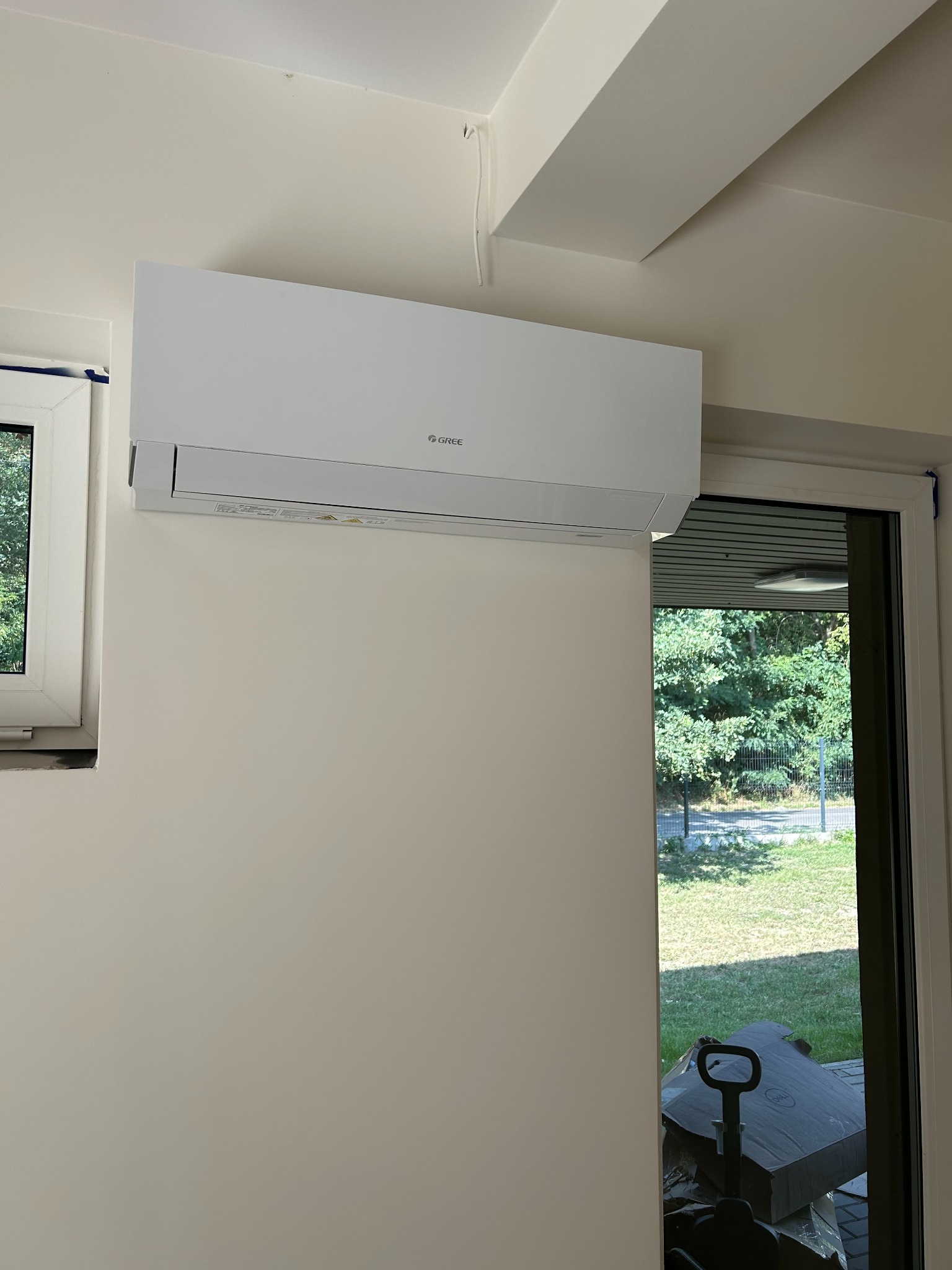 Biały klimatyzator marki Gree zamontowany na ścianie w pomieszczeniu, widoczne okno z niebieską taśmą malarską oraz przesuwne drzwi balkonowe z widokiem na zieleń i kartony w tle.