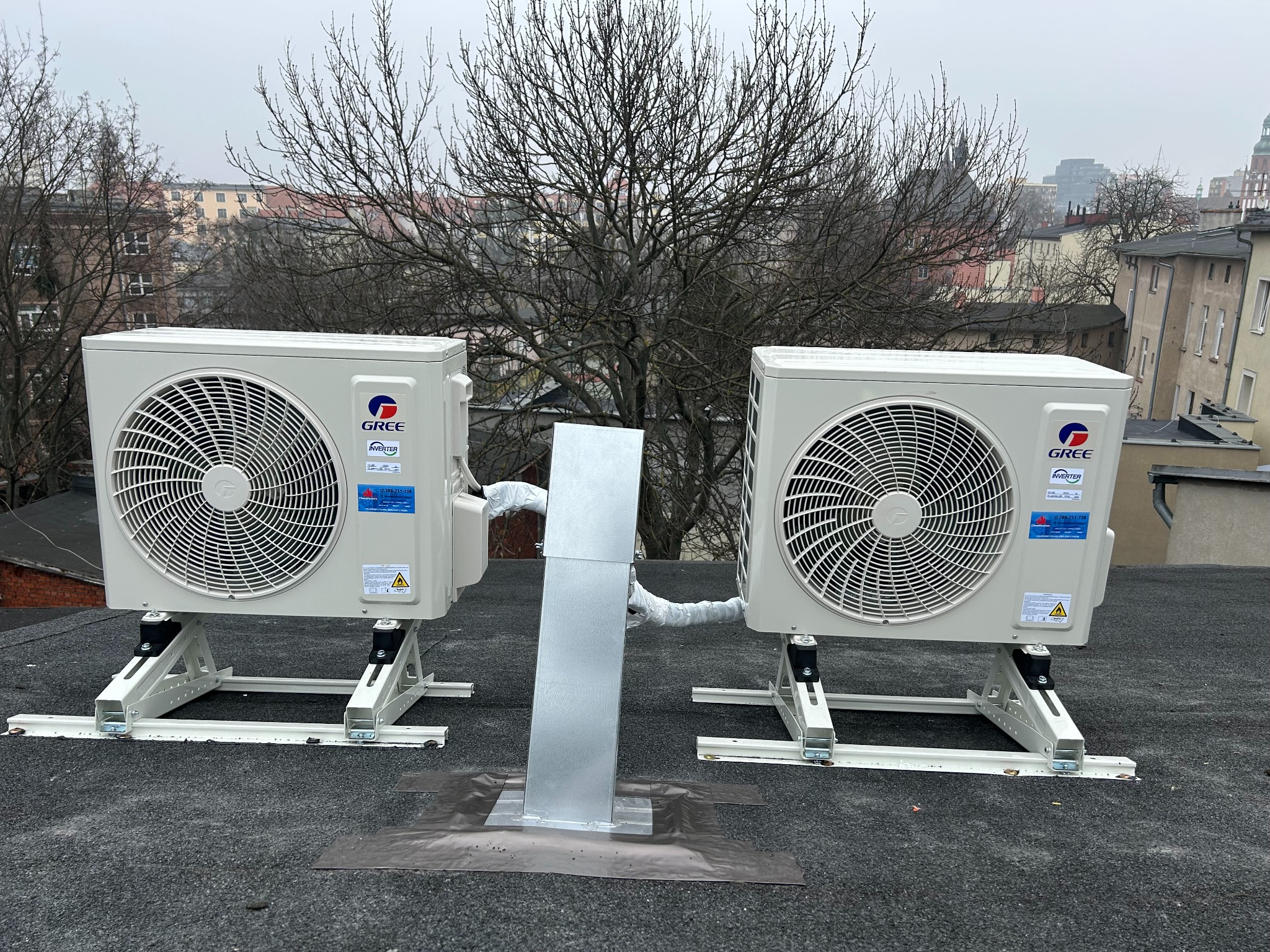Dwa klimatyzatory marki Gree zamontowane na dachu budynku z widoczną instalacją odprowadzającą skropliny, na tle zabudowy miejskiej i drzew bez liści.