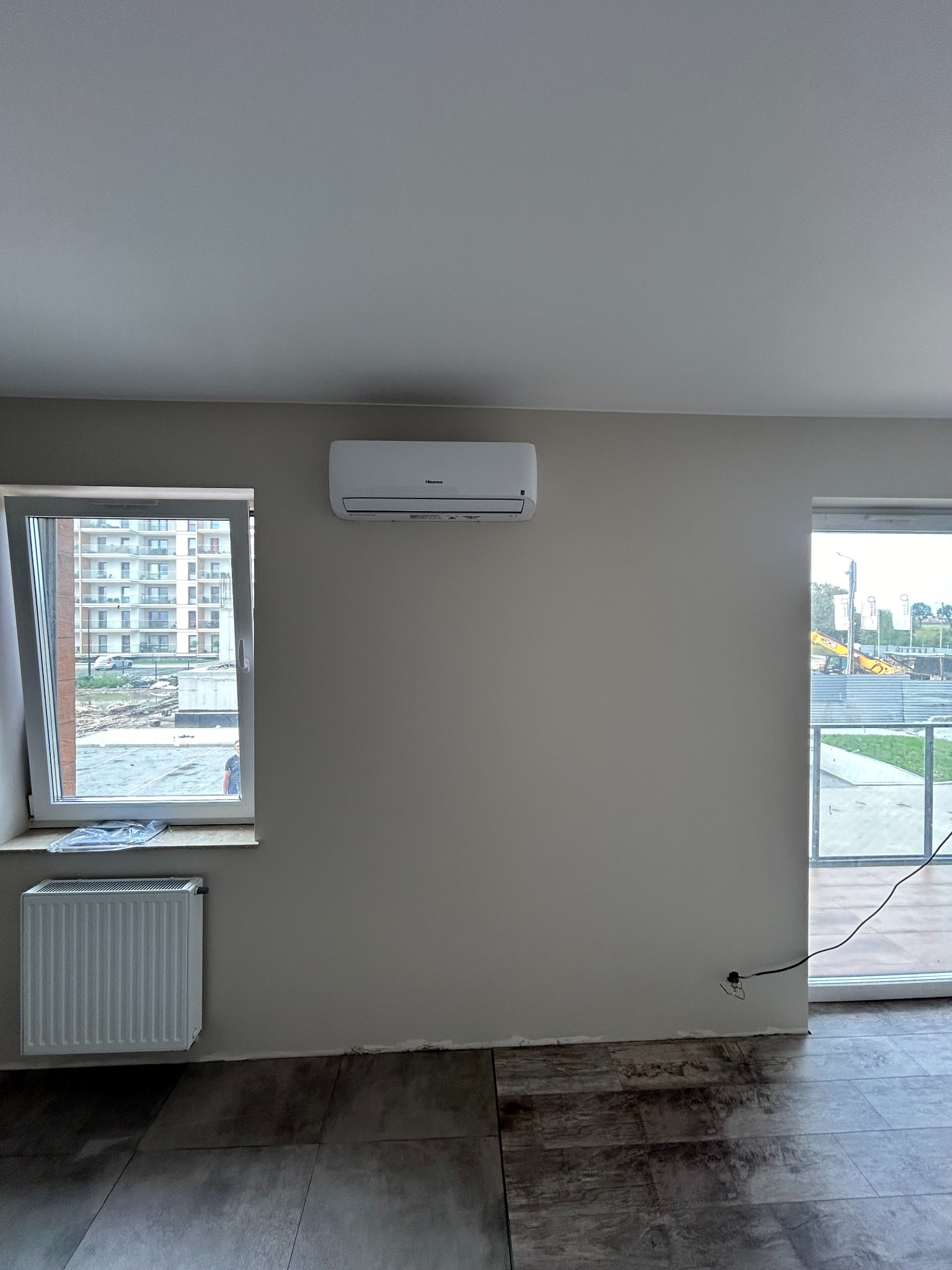 Biały klimatyzator Hisense zamontowany na szarej ścianie w surowym wnętrzu, widok przez okno na budowę i blok mieszkalny, grzejnik pod oknem, ciemne płytki podłogowe.