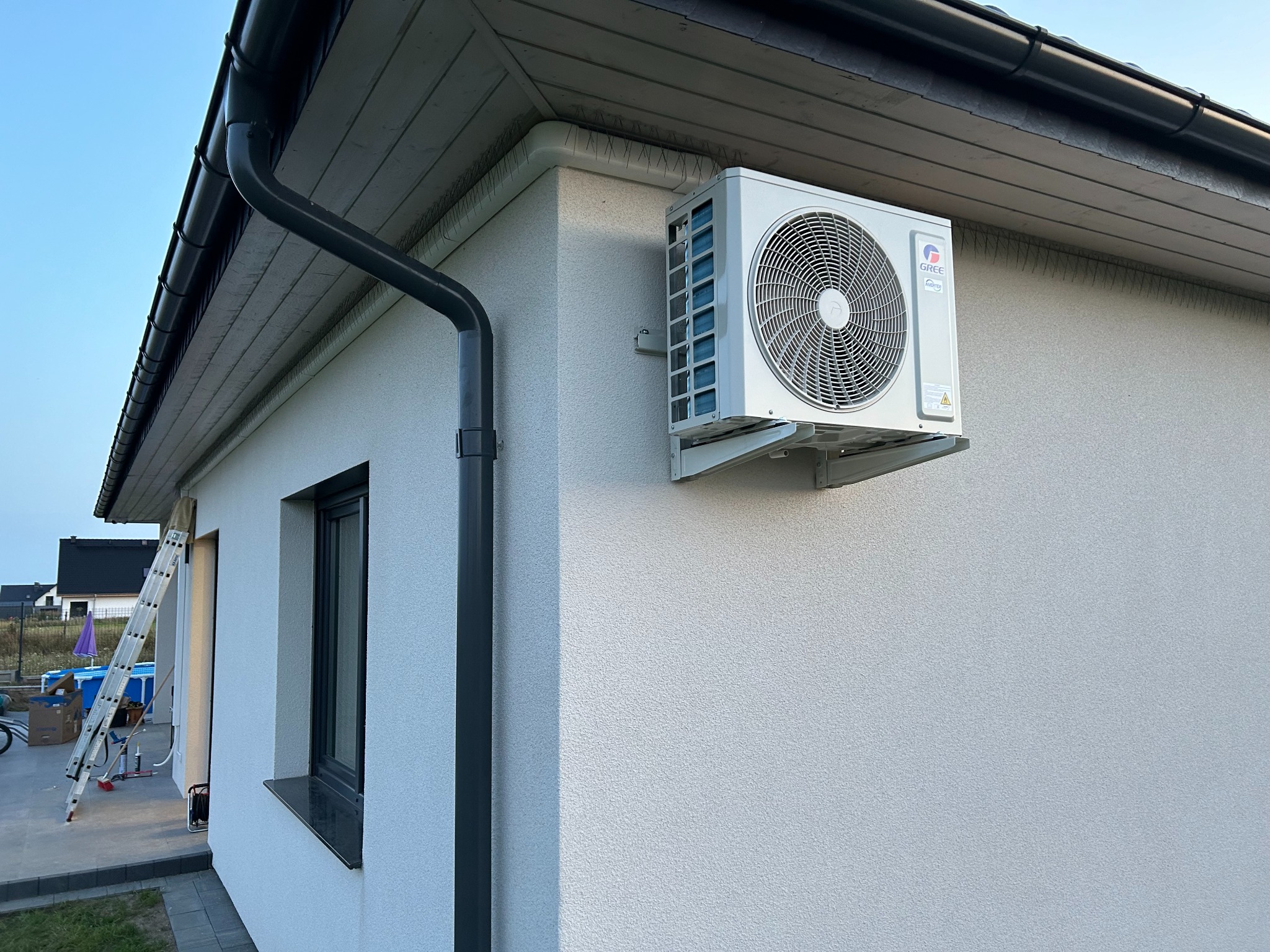 Biała jednostka zewnętrzna klimatyzacji Gree zamontowana na ścianie domu z szarym orynnowaniem, widoczna drabina i basen w tle.