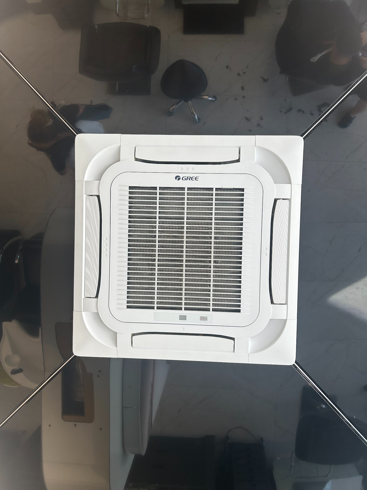 Sufitowa kaseta klimatyzacyjna marki Gree w pomieszczeniu biurowym z ciemną podłogą i meblami, widok z dołu.