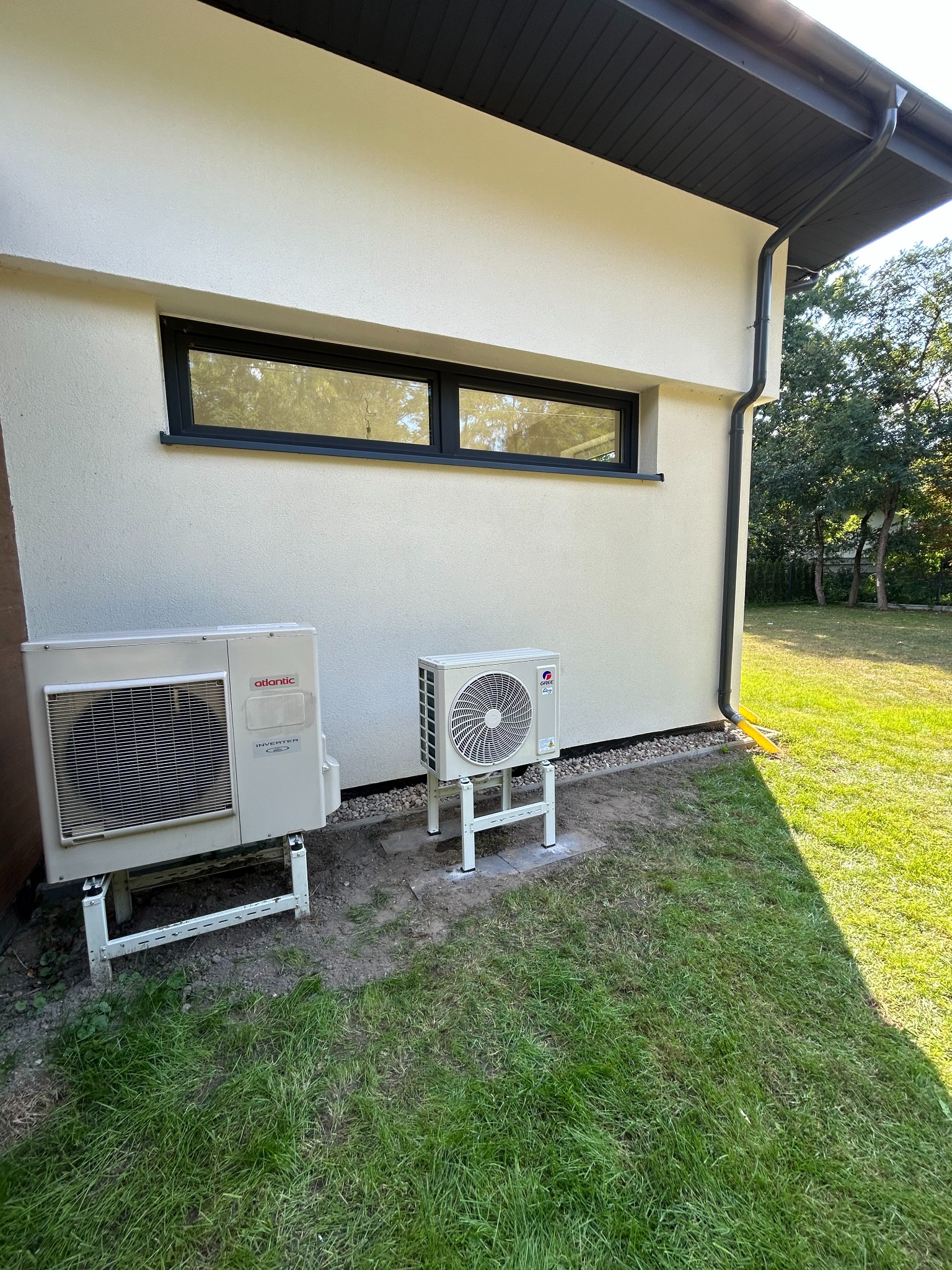 Dwa zewnętrzne agregaty klimatyzacji (Atlantic i Gree) zamontowane na metalowych wspornikach przy ścianie domu z oknem i czarną rynną, widok z trawnika.