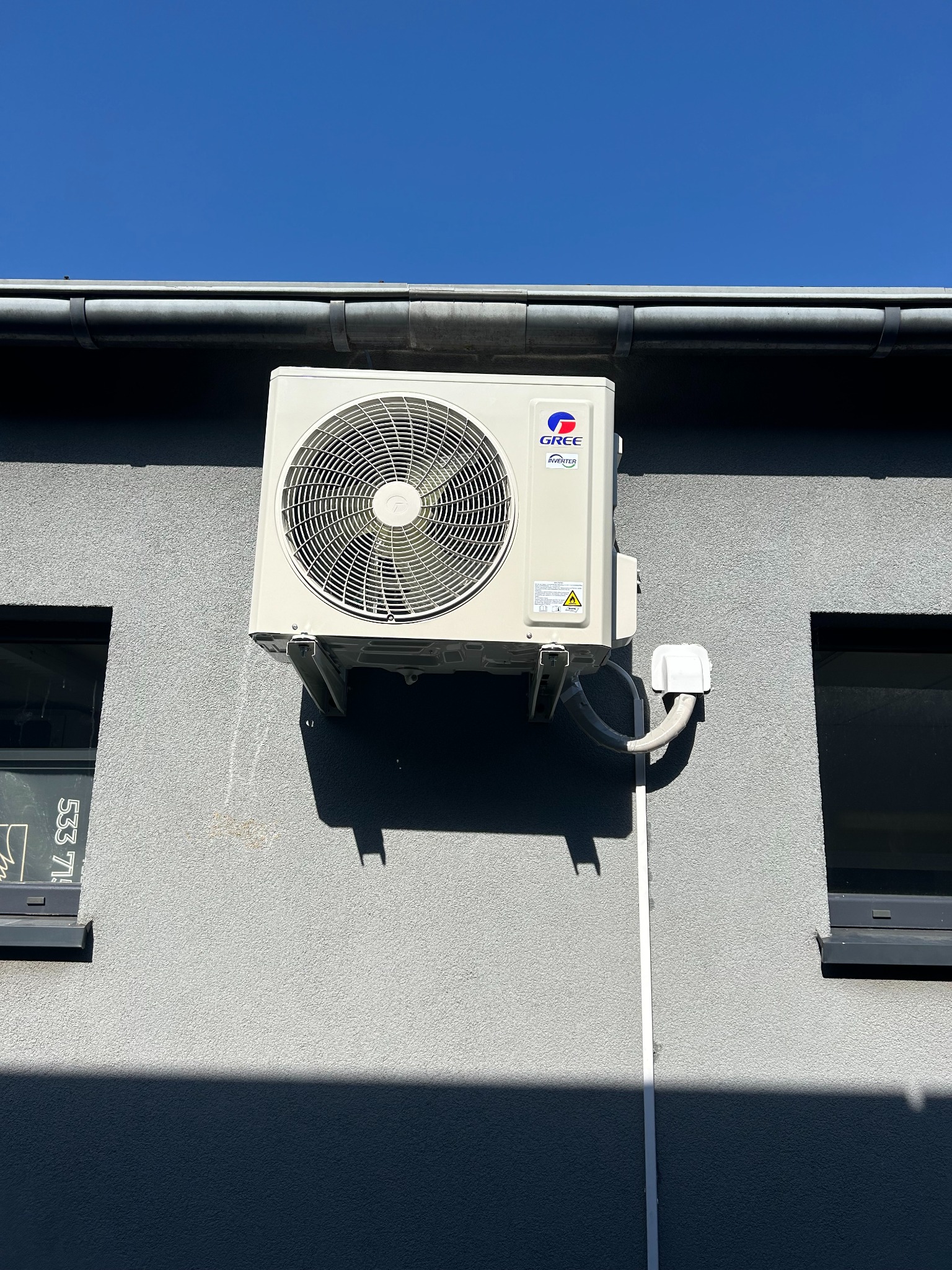 Zewnętrzna jednostka klimatyzacji marki Gree zamontowana na szarej elewacji budynku, widoczne dwa okna i rynna, błękitne niebo w tle.