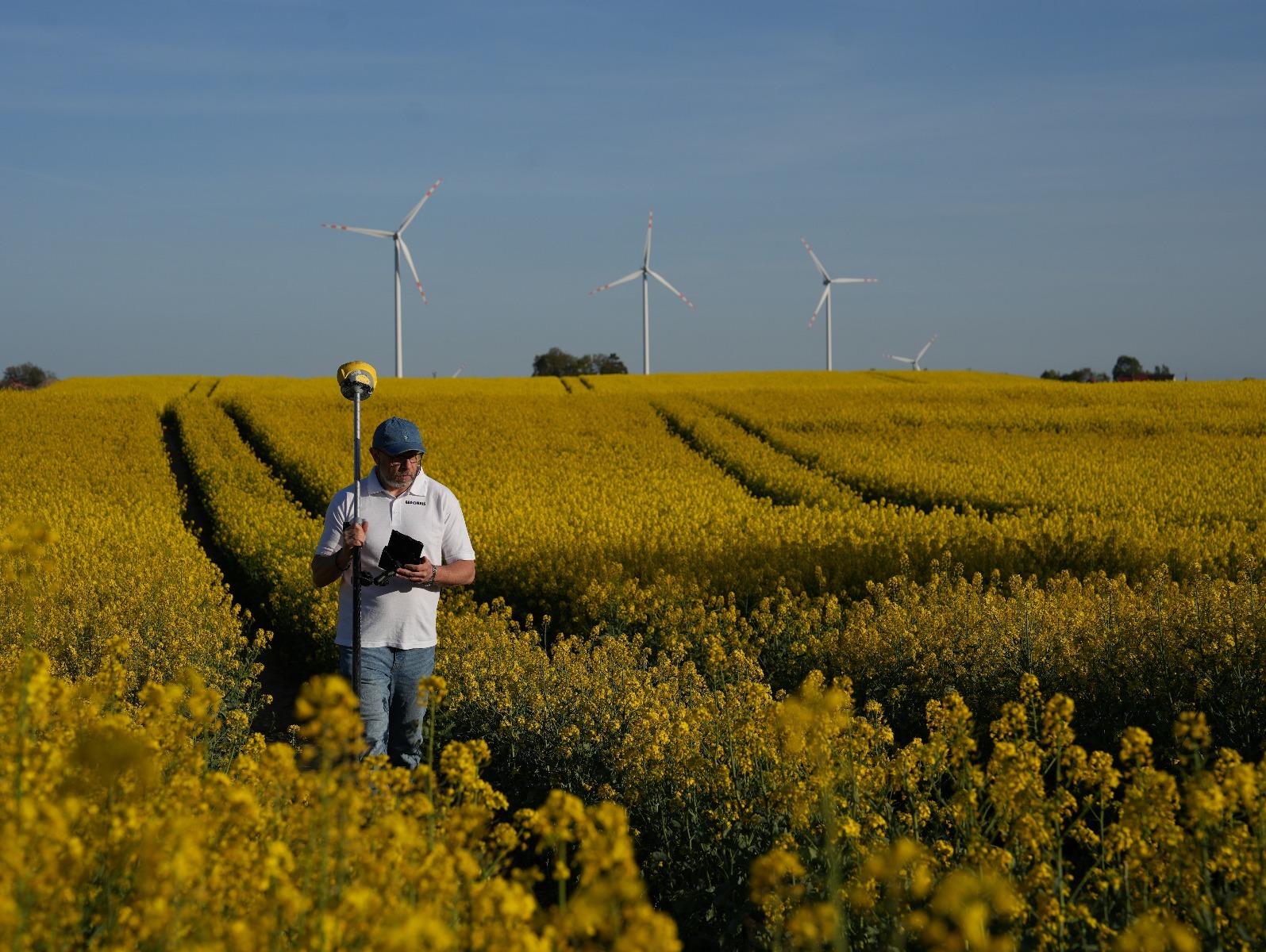 Mężczyzna z odbiornikiem GPS w polu rzepaku, w tle turbiny wiatrowe. Pomiar terenu z wykorzystaniem nowoczesnych technologii.