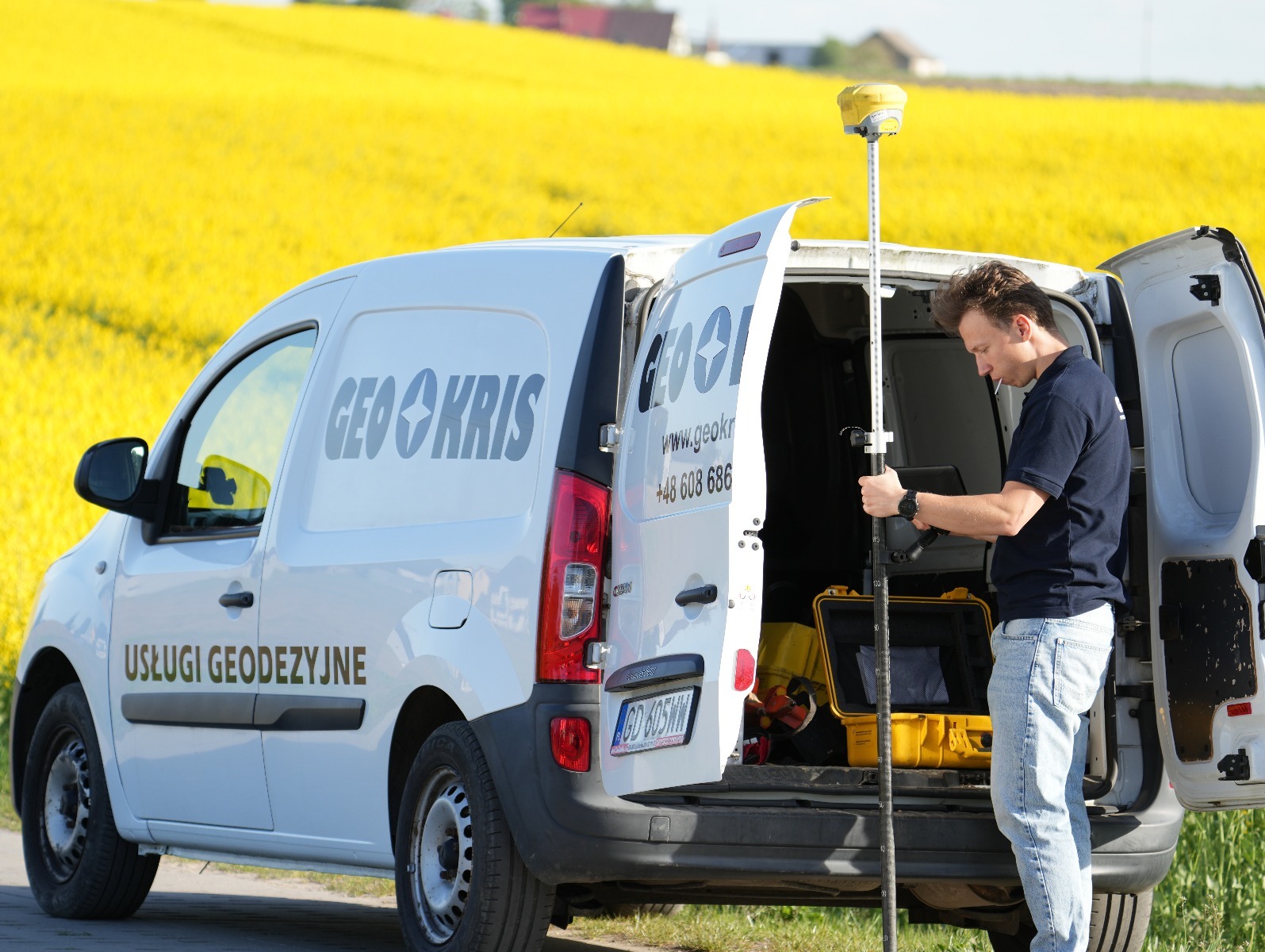 Pracownik firmy GEO-KRIS obsługuje precyzyjny sprzęt pomiarowy na tle kwitnącego pola rzepaku. Biały bus z otwartymi drzwiami, żółta walizka ze sprzętem.