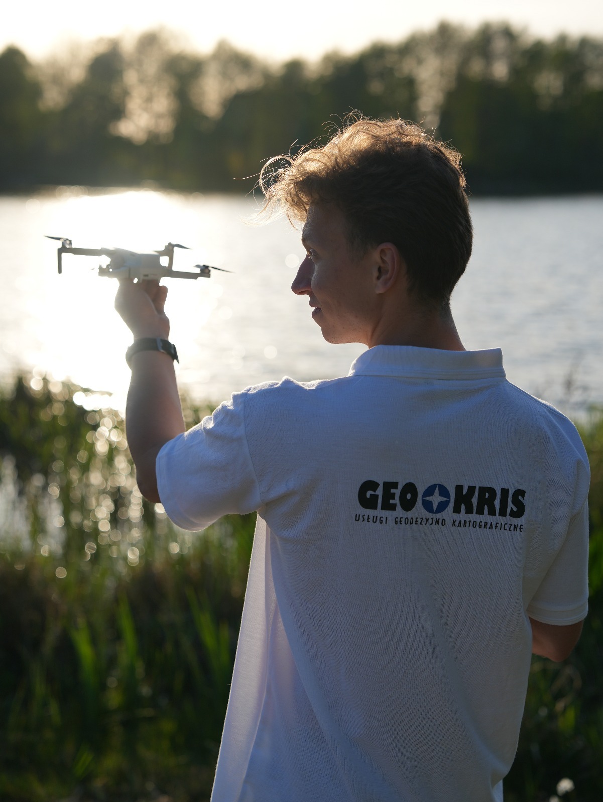 Młody mężczyzna z dronem na tle jeziora, na koszulce logo firmy GEOKRIS - Usługi Geodezyjno Kartograficzne. Ujęcie z perspektywy osoby trzeciej.