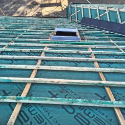 ERMA BUD SPÓŁKA Z OGRANICZONĄ ODPOWIEDZIALNOŚCIĄ - Konstrukcja dachu z oknem dachowym w trakcie montażu, widok z góry. Zielona membrana dachowa z czarnym nadrukiem, drewniane łaty. Fragment buta w dolnej części kadru.