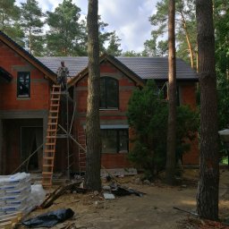 ERMA BUD SPÓŁKA Z OGRANICZONĄ ODPOWIEDZIALNOŚCIĄ - Budowa domu z cegły w Warszawie, widok na częściowo pokryty dach z drabiny i rusztowania. W tle las.