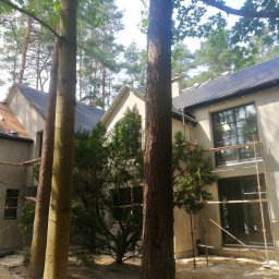 ERMA BUD SPÓŁKA Z OGRANICZONĄ ODPOWIEDZIALNOŚCIĄ - Nowoczesny dom z ciemną dachówką i jasną elewacją w trakcie prac wykończeniowych. Widoczne rusztowania i drzewa w otoczeniu.