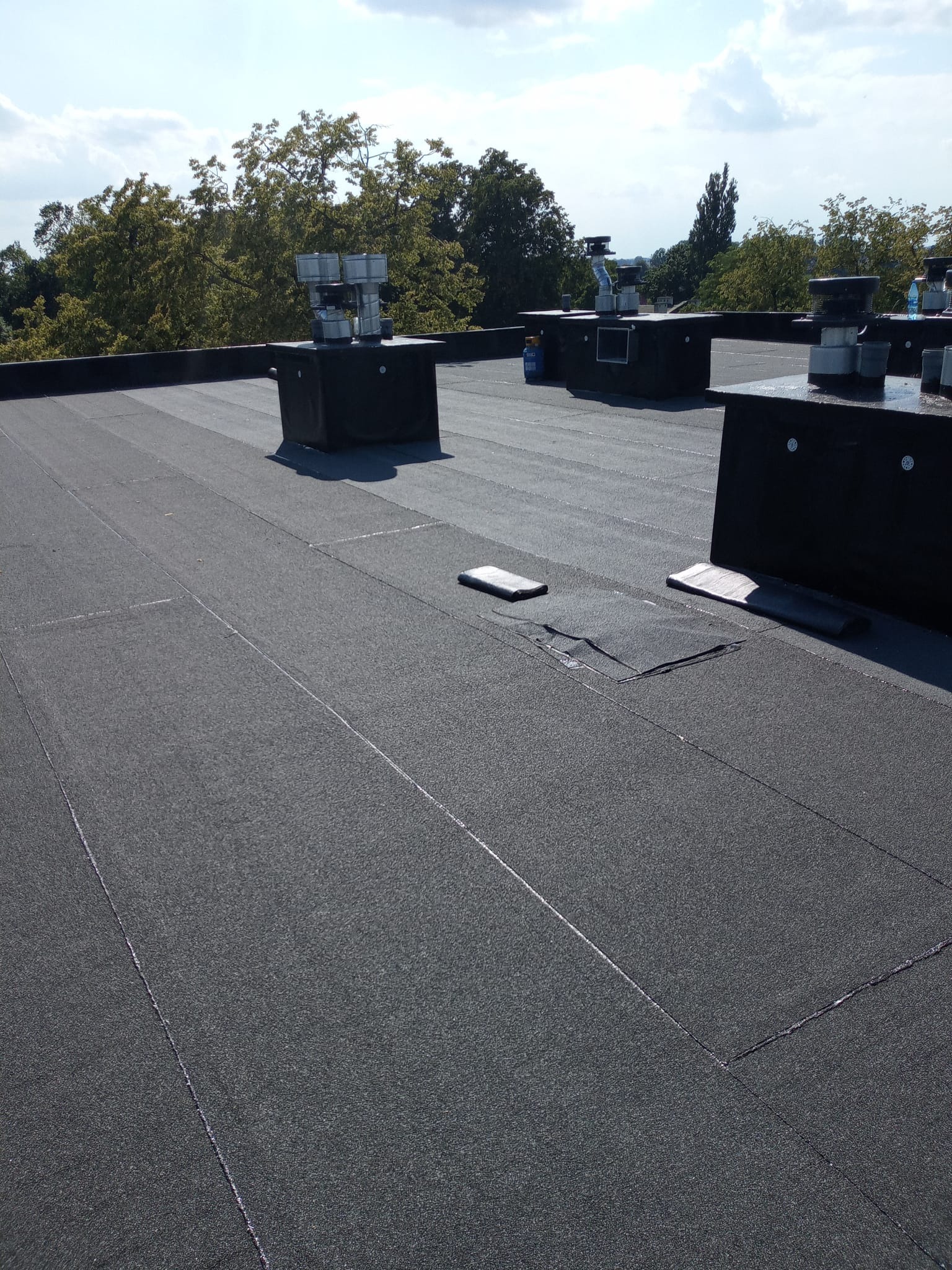 Czarny, płaski dach pokryty papą termozgrzewalną z kominami wentylacyjnymi. Widoczne łączenia papy i elementy obróbek. W tle zieleń drzew i błękitne niebo.