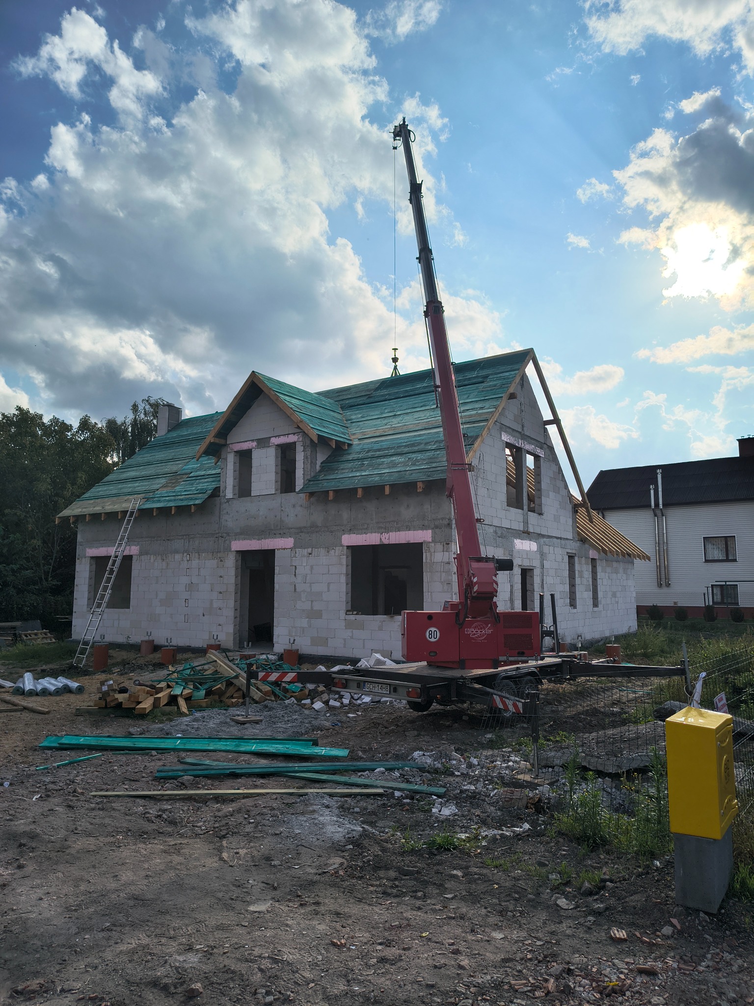 Budowa domu jednorodzinnego w trakcie realizacji. Widoczny dźwig, częściowo pokryty dach z zieloną membraną, elementy więźby dachowej. Na pierwszym planie materiały budowlane.