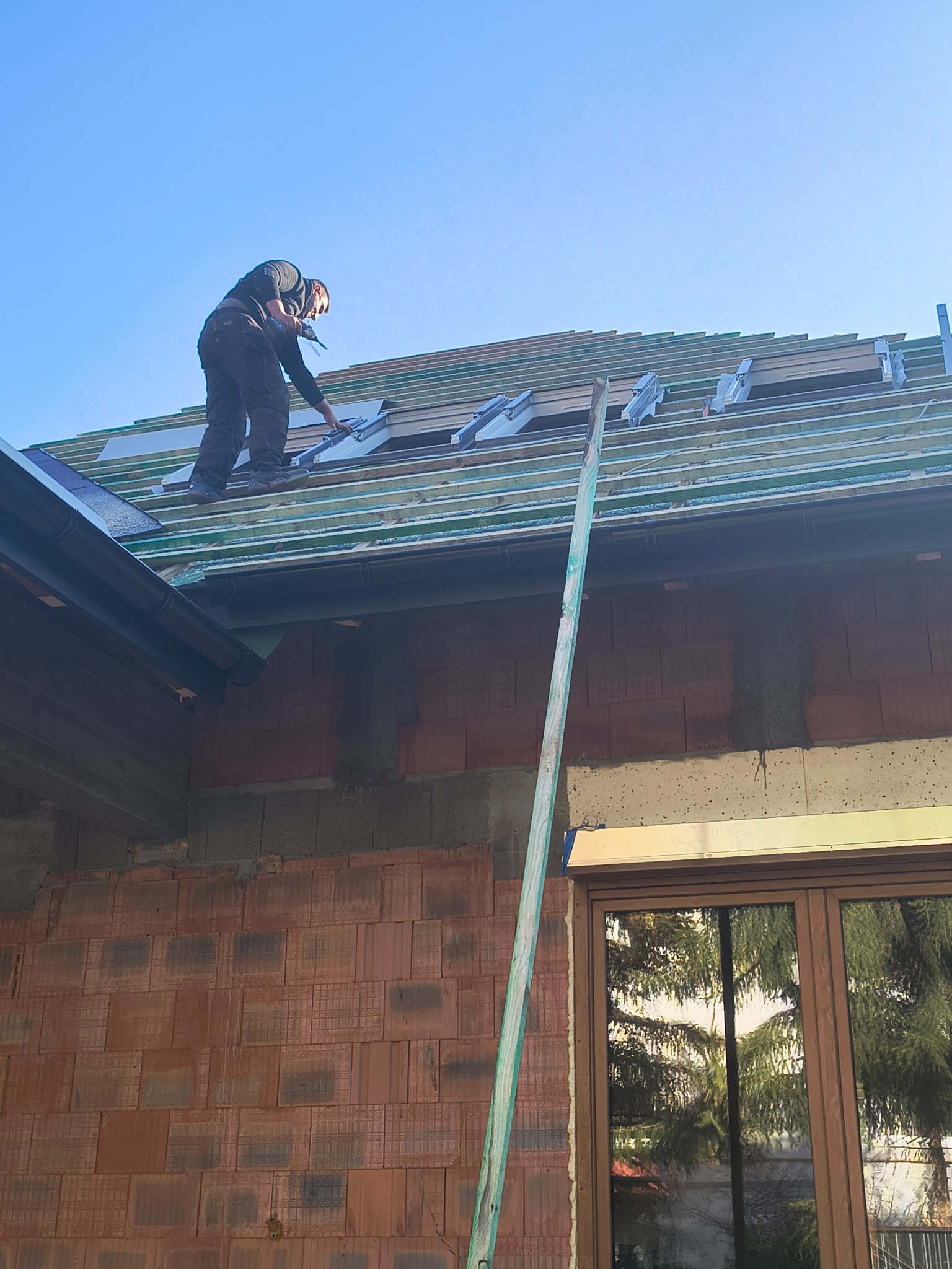 Praca na dachu w słoneczny dzień: dekarz montuje elementy konstrukcyjne na drewnianej więźbie, widok z dołu na budynek w trakcie budowy.