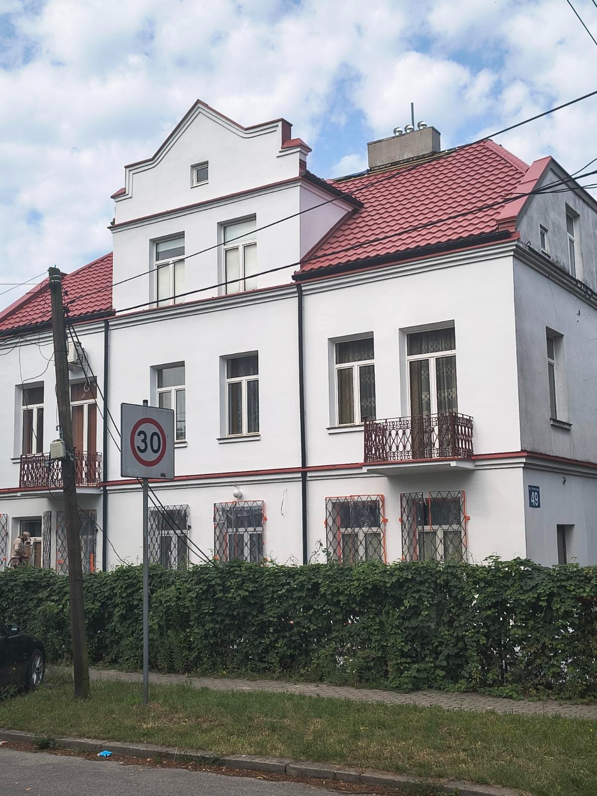 Biały budynek mieszkalny z czerwoną dachówką, widoczny z poziomu ulicy, z zielonym żywopłotem i znakiem drogowym. Uwagę przykuwa odnowiona elewacja i symetryczny układ okien.