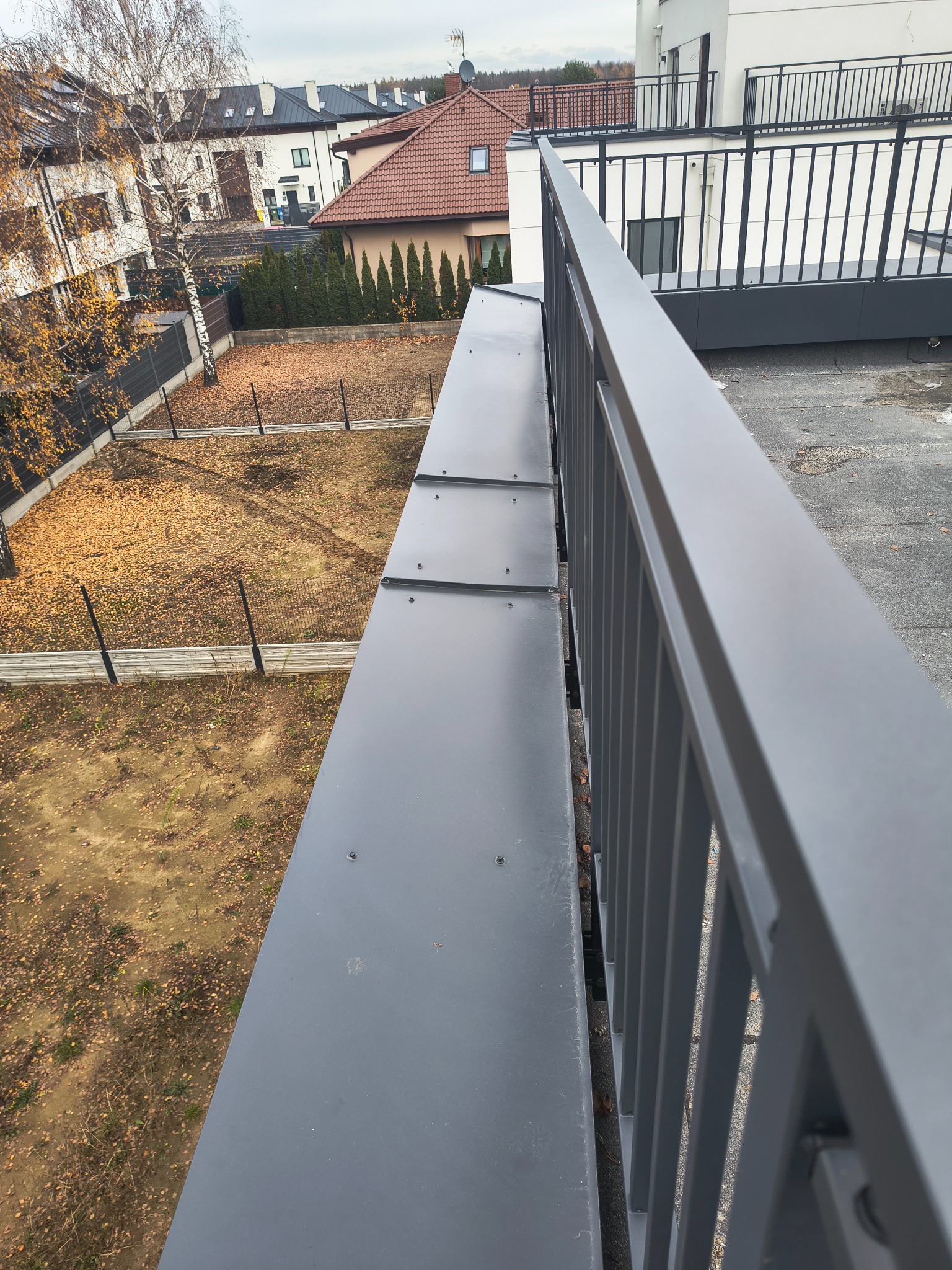 Szare obróbki blacharskie na dachu z widokiem na jesienny ogród i zabudowę jednorodzinną w tle. Ujęcie z góry, z bliska na detale.