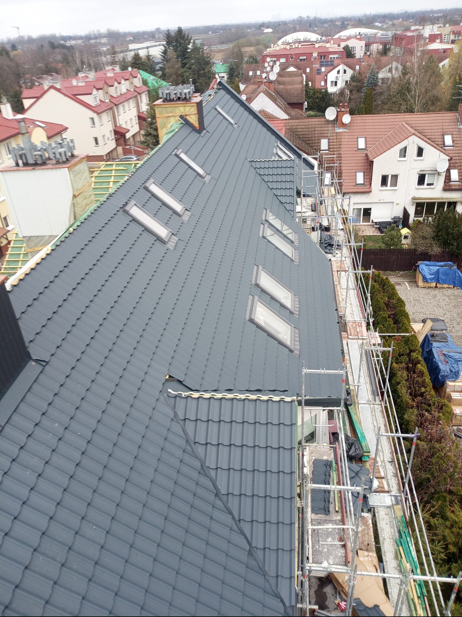 Szary dach z oknami dachowymi w trakcie montażu, widoczne rusztowania i otoczenie budynków mieszkalnych. Ujęcie z góry, pochmurne niebo.
