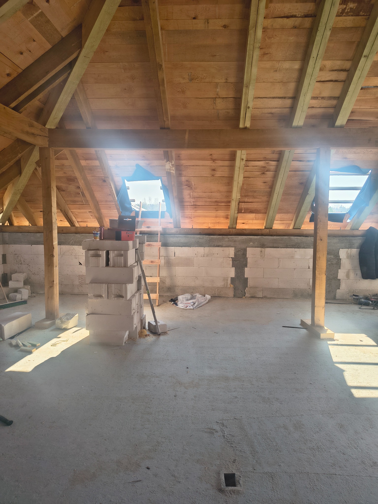 Surowe poddasze z drewnianą konstrukcją dachu i częściowo wymurowanymi ścianami z bloczków. Widoczne materiały budowlane i drabina. Wpadające światło słoneczne.