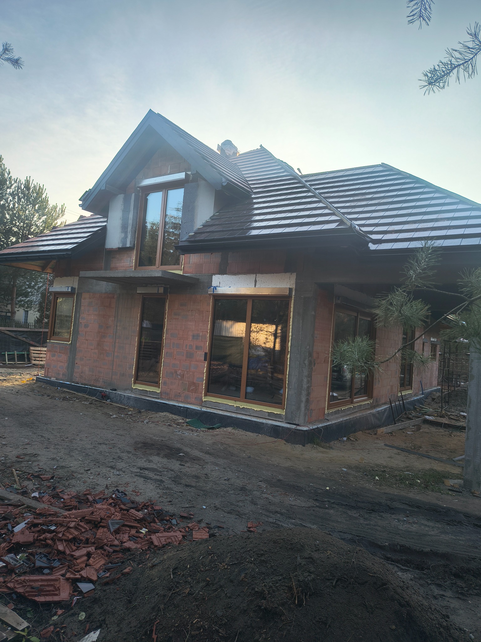 Dom w trakcie budowy z częściowo ukończonym dachem z brązowej dachówki. Widoczne okna i ściany z czerwonej cegły. Na pierwszym planie gruz i ziemia.