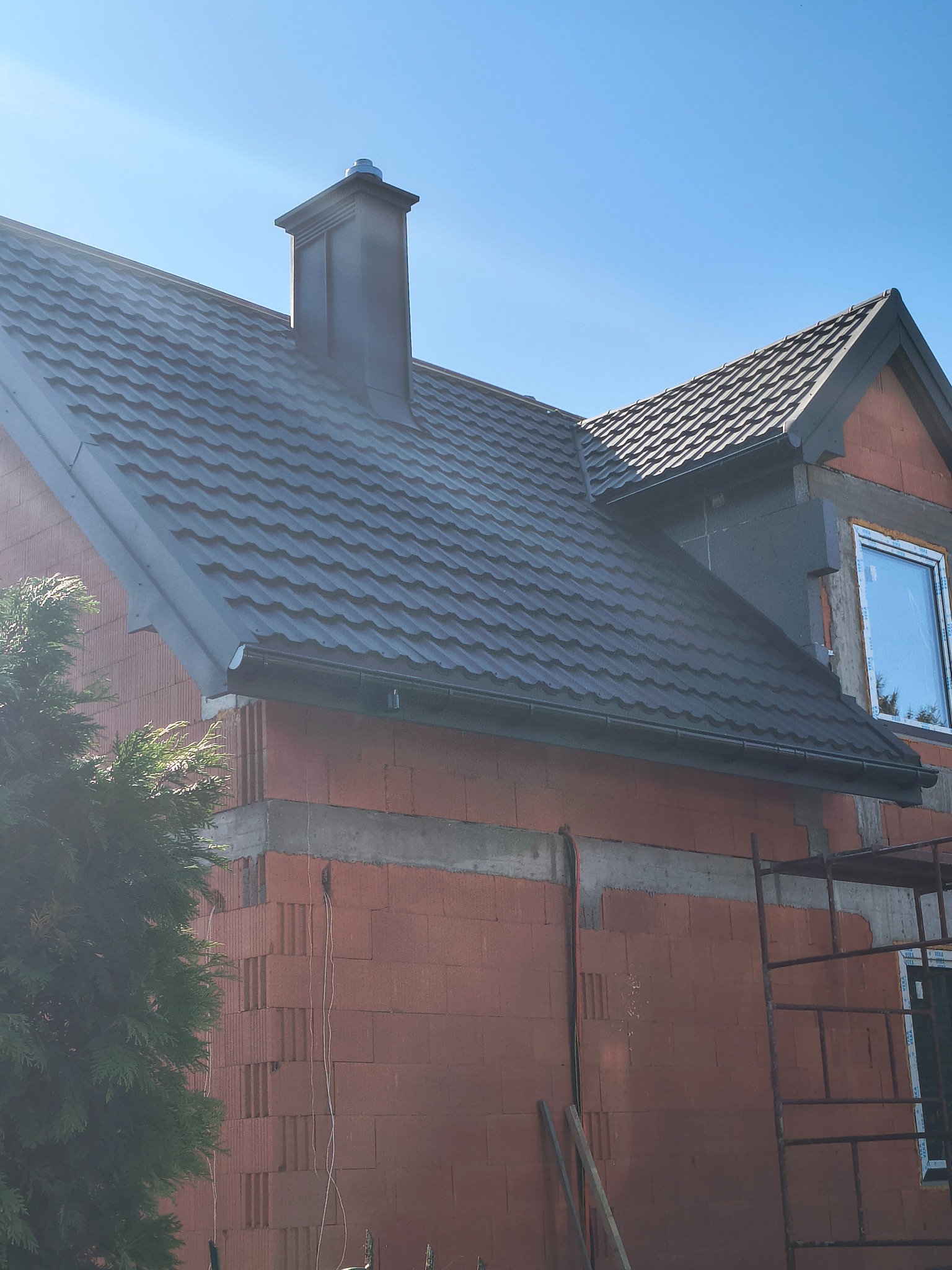 Nowy, grafitowy dach z blachodachówki modułowej, komin w kolorze dachu, orynnowanie. Budynek w trakcie budowy, widoczne rusztowanie i niezakończona elewacja.