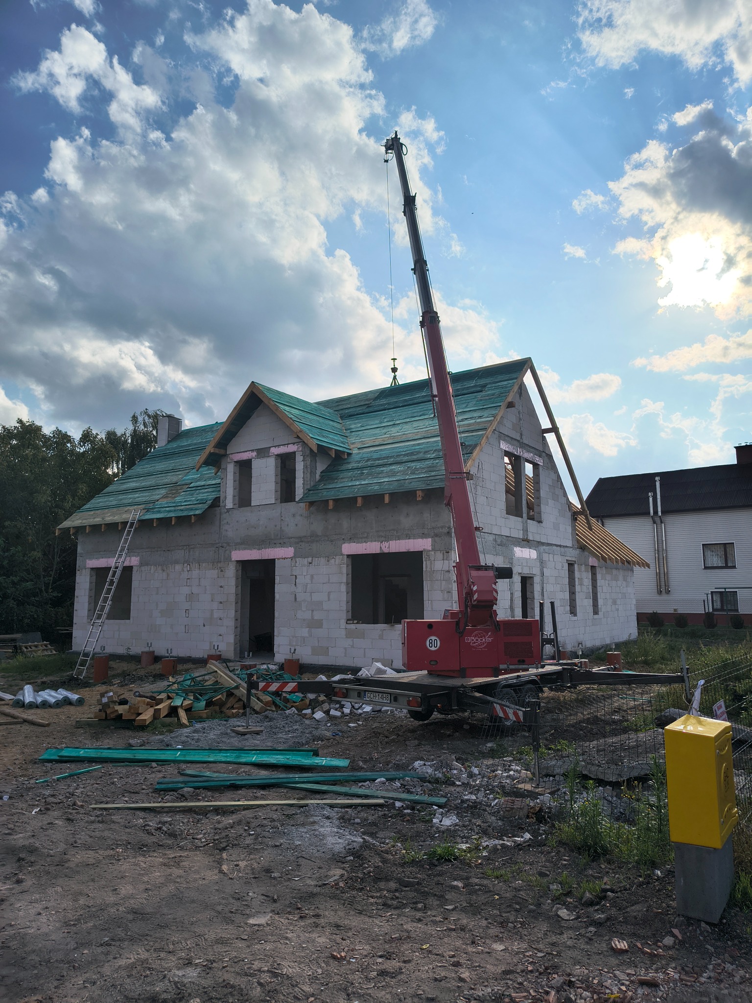 Budowa domu z użyciem dźwigu: zielone deski na dachu, białe ściany z pustaków, widoczne drewno i drabina. Słoneczny dzień, częściowo zachmurzone niebo.