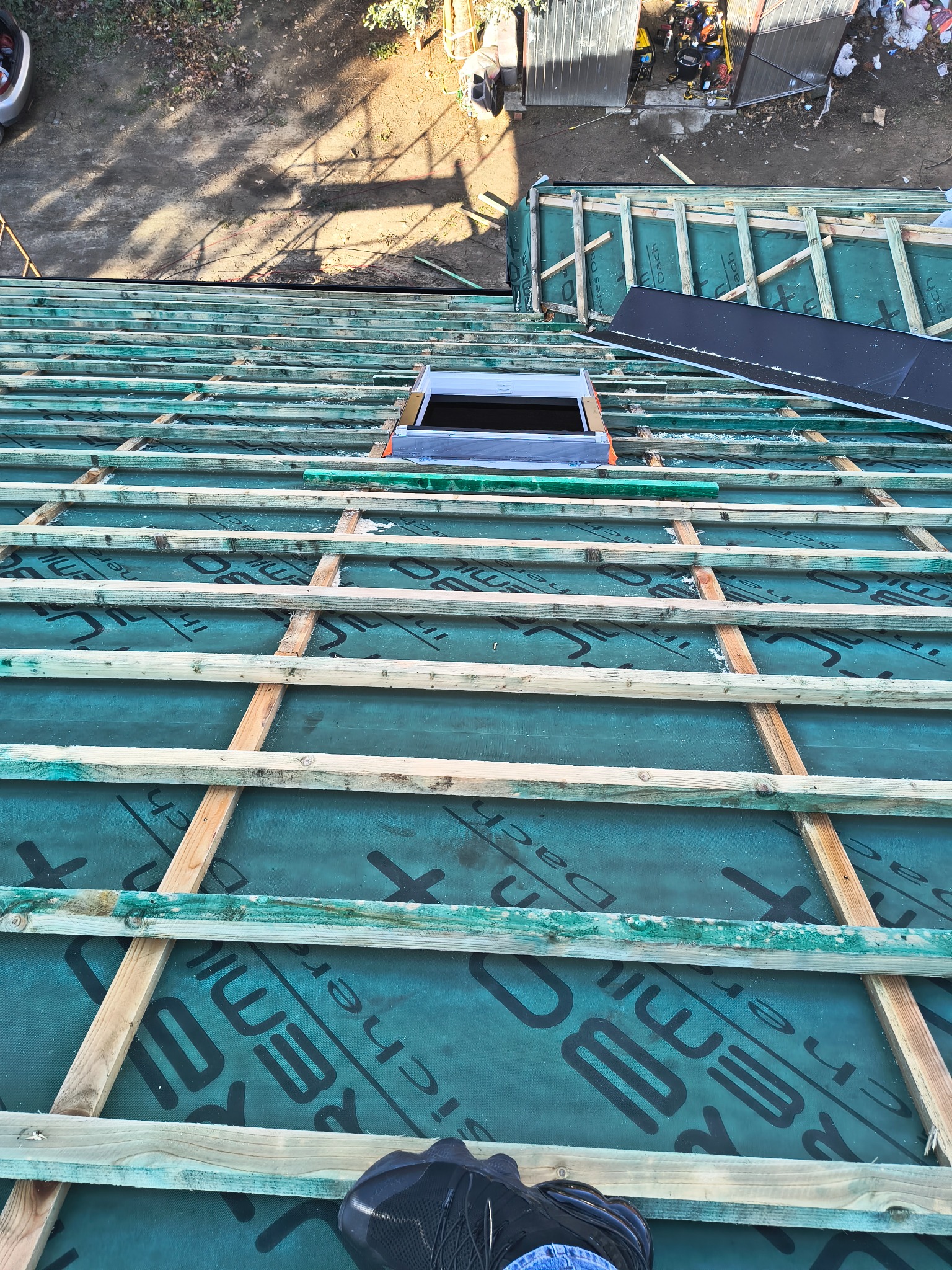 Konstrukcja dachu z oknem dachowym w trakcie montażu, widok z góry. Zielona membrana dachowa z czarnym nadrukiem, drewniane łaty. Fragment buta w dolnej części kadru.