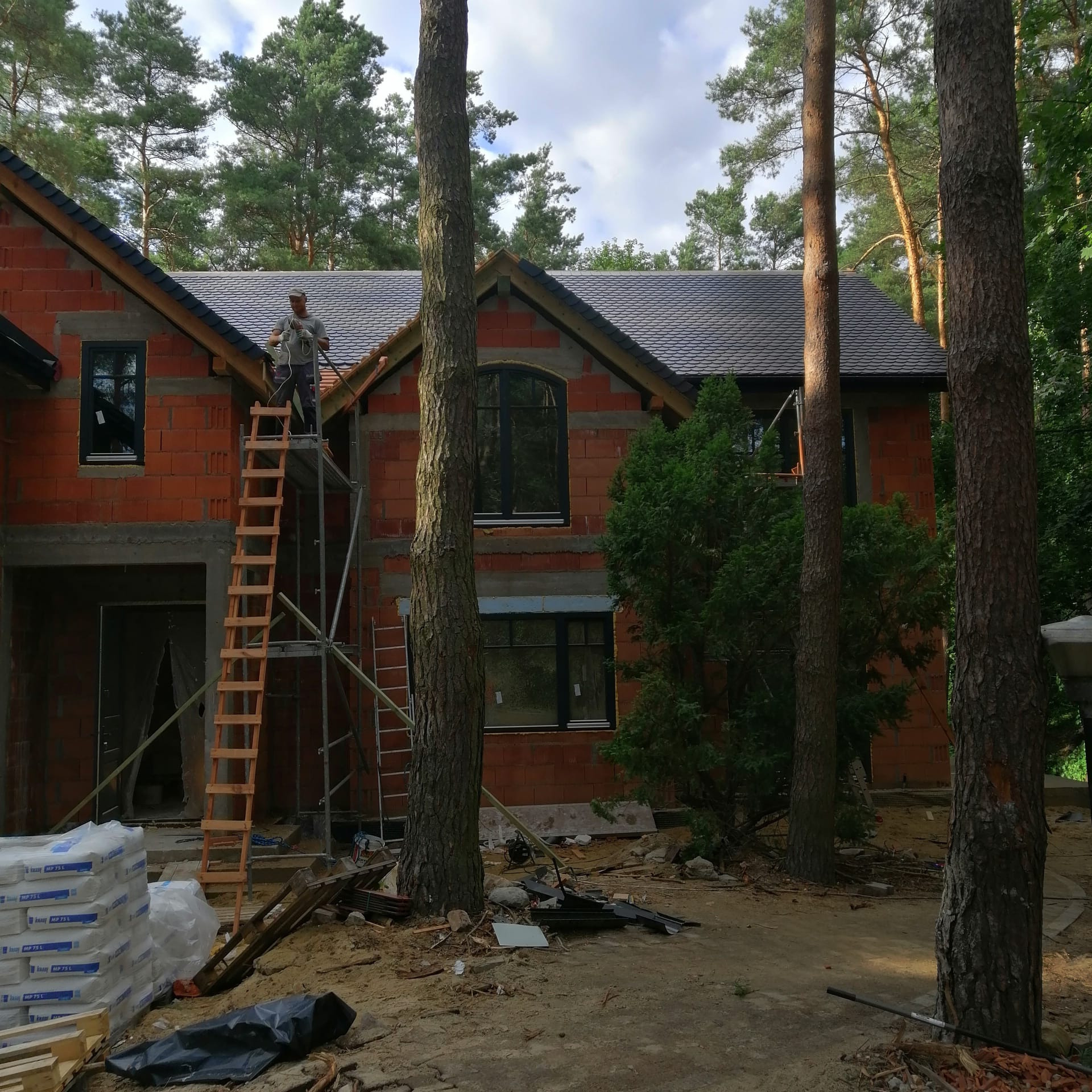 Budowa domu z cegły w Warszawie, widok na częściowo pokryty dach z drabiny i rusztowania. W tle las.