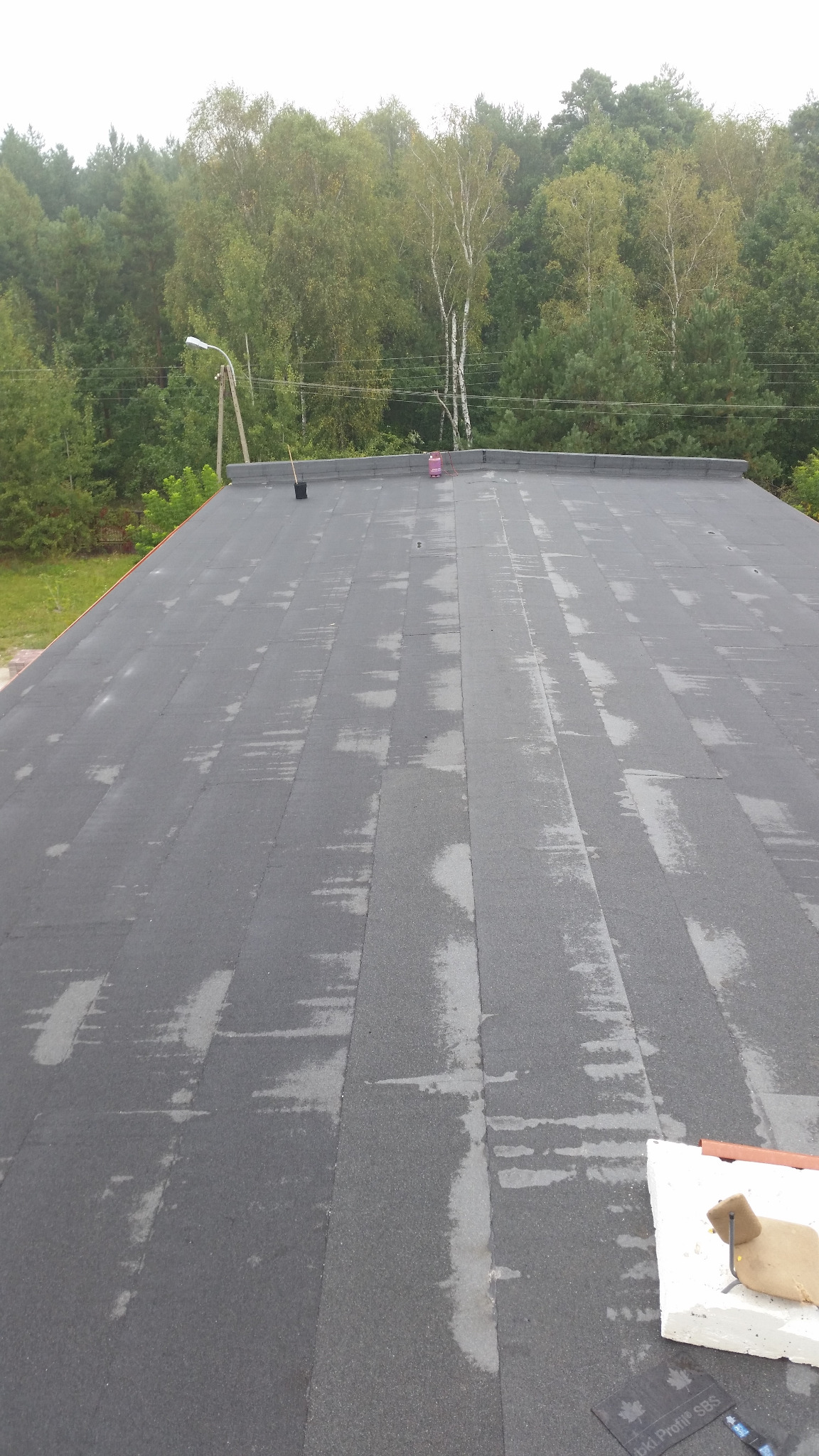 Widok z góry na płaski, pokryty papą dach z widocznymi śladami wilgoci. W tle drzewa i słup oświetleniowy. Na dachu leżą materiały budowlane.