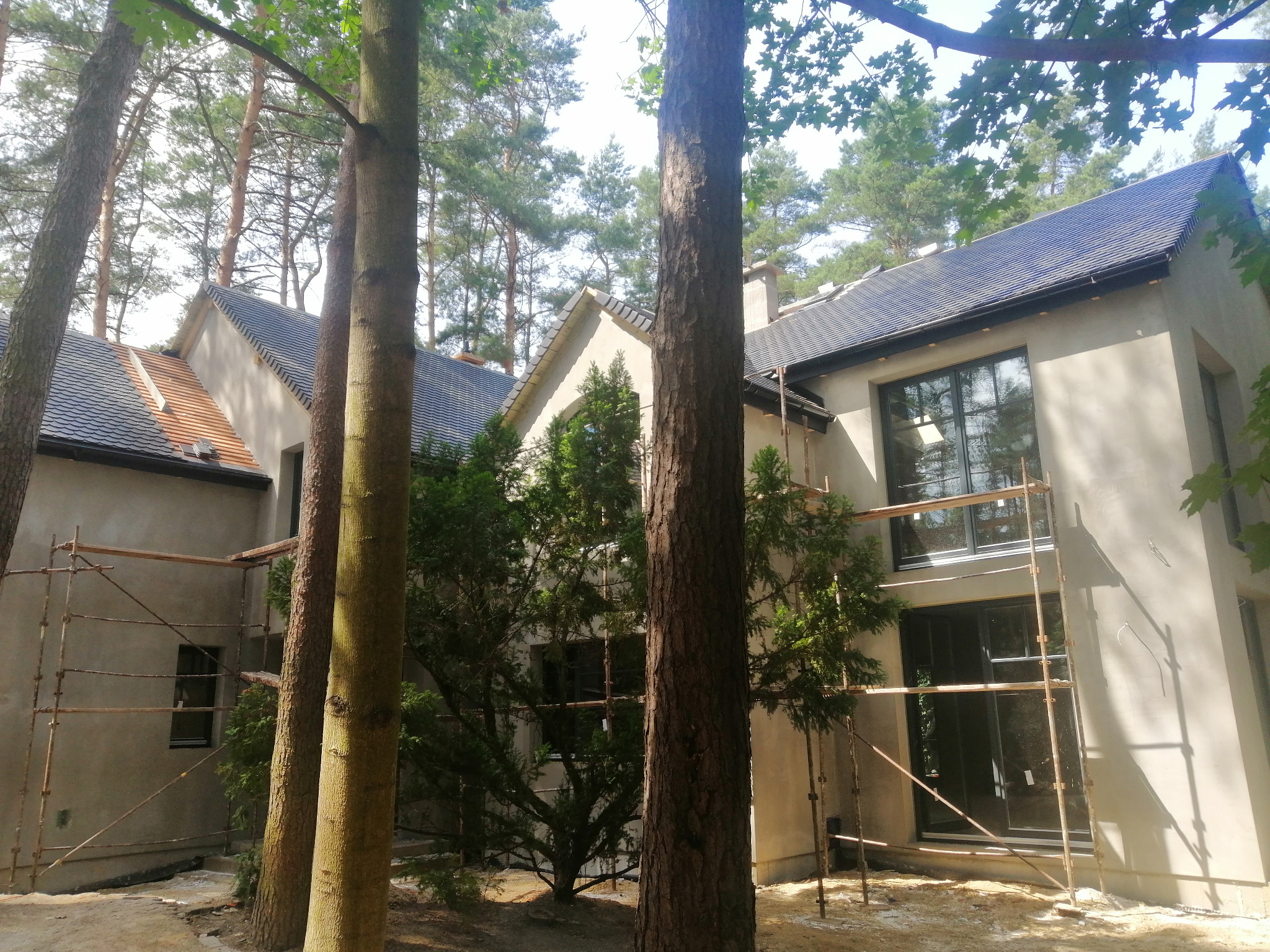 Nowoczesny dom z ciemną dachówką i jasną elewacją w trakcie prac wykończeniowych. Widoczne rusztowania i drzewa w otoczeniu.