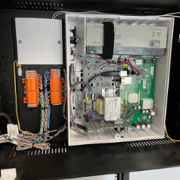 Otwarta szafka rack z centralą alarmową Jablotron JA-100K, widoczne okablowanie, moduły z pomarańczowymi złączkami i płyta główna z elektroniką.