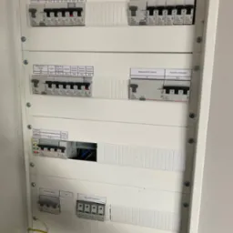 Tablica rozdzielcza z bezpiecznikami automatycznymi, widoczne wyłączniki różnicowoprądowe, ograniczniki przepięć i rozłącznik izolacyjny, opisane obwody zasilające.