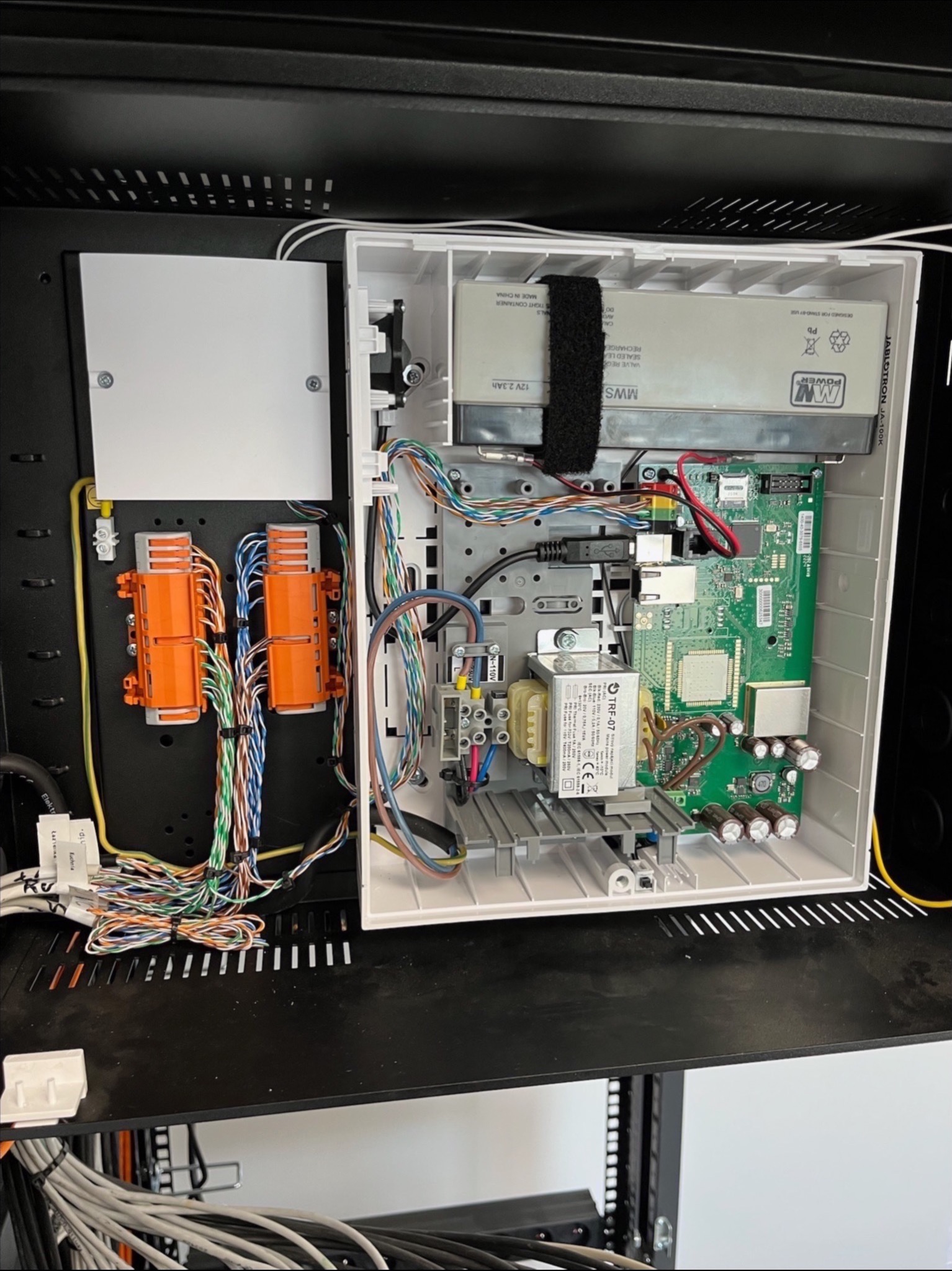 Otwarta szafka rack z centralą alarmową Jablotron JA-100K, widoczne okablowanie, moduły z pomarańczowymi złączkami i płyta główna z elektroniką.