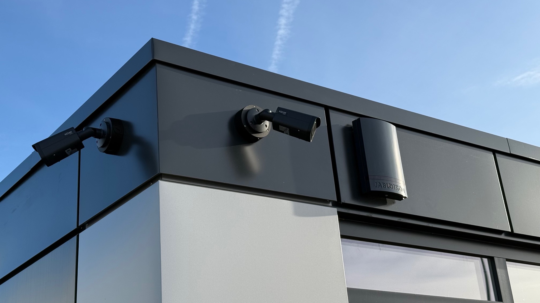 Dwie kamery monitoringu marki KOZAK i moduł Jablotron zamontowane na elewacji budynku o nowoczesnym designie, widok z dołu na błękitne niebo.