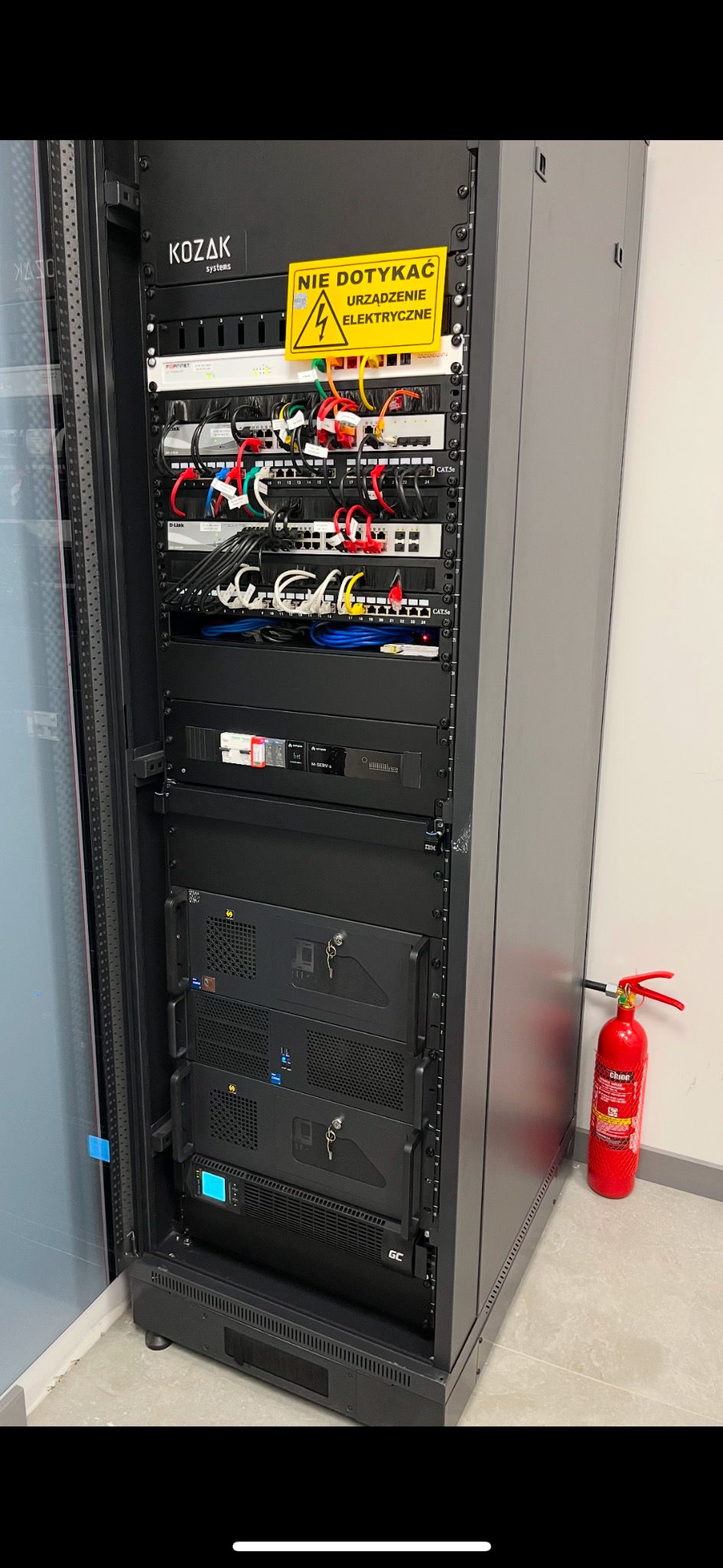 Czarna szafa rack z zainstalowanymi urządzeniami sieciowymi i zasilającymi, ostrzeżenie 'Nie dotykać - Urządzenie elektryczne' na górze, czerwona gaśnica obok.
