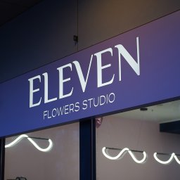 Kluma Studio - Podświetlany szyld z nazwą 'Eleven Flowers Studio' na fioletowym tle, wykonany z dibondu i folii, z widocznym ciepłym światłem LED. W tle widoczne wnętrze kwiaciarni z dekoracjami i osobą.