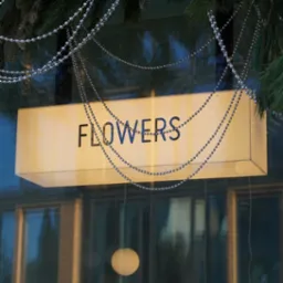 Świąteczna dekoracja witryny kwiaciarni z podświetlanym szyldem 'FLOWERS', ozdobiona girlandami z koralików i sztucznymi sopelkami, odbicie wnętrza sklepu w szybie.