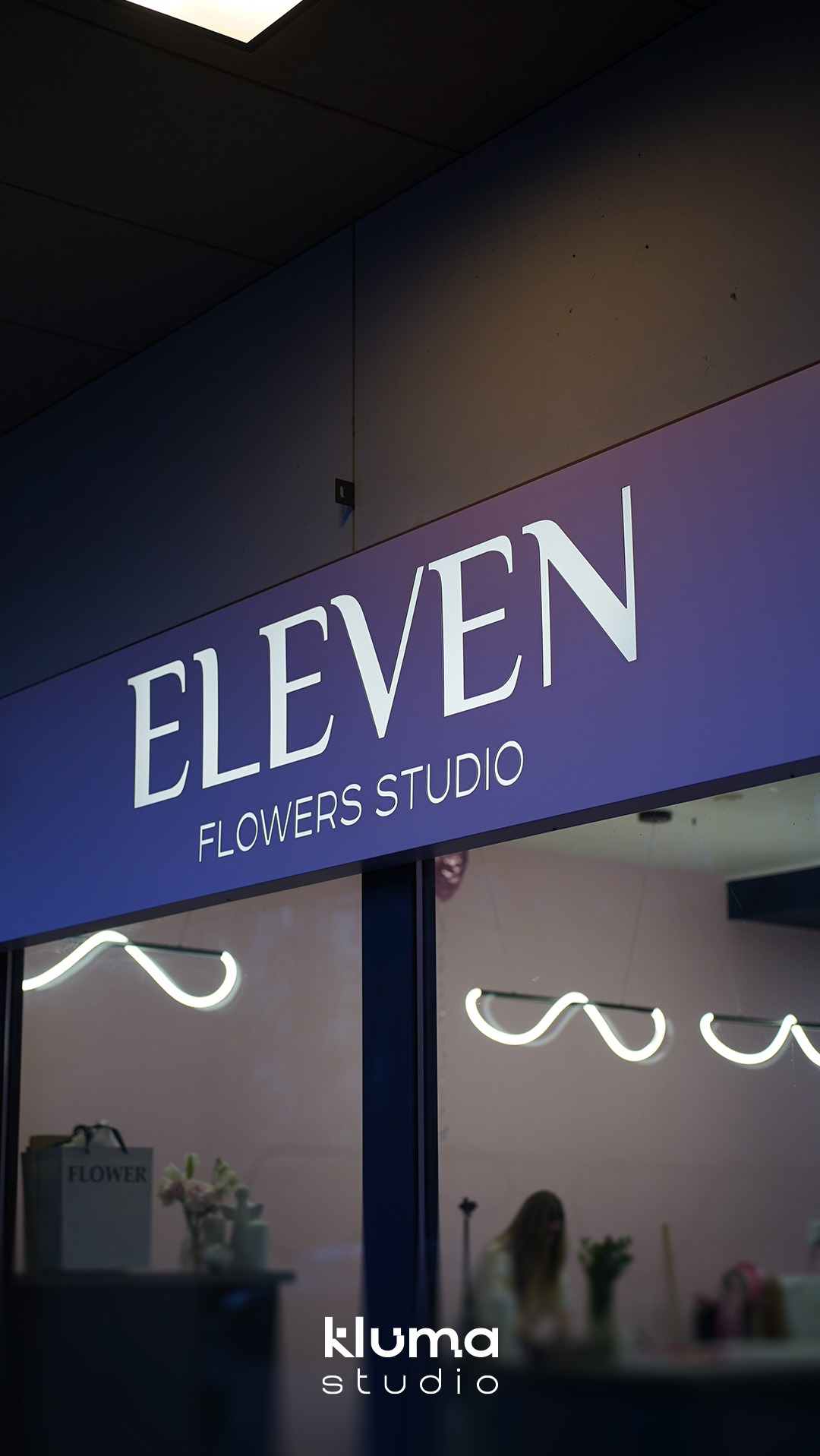 Podświetlany szyld z nazwą 'Eleven Flowers Studio' na fioletowym tle, wykonany z dibondu i folii, z widocznym ciepłym światłem LED. W tle widoczne wnętrze kwiaciarni z dekoracjami i osobą.