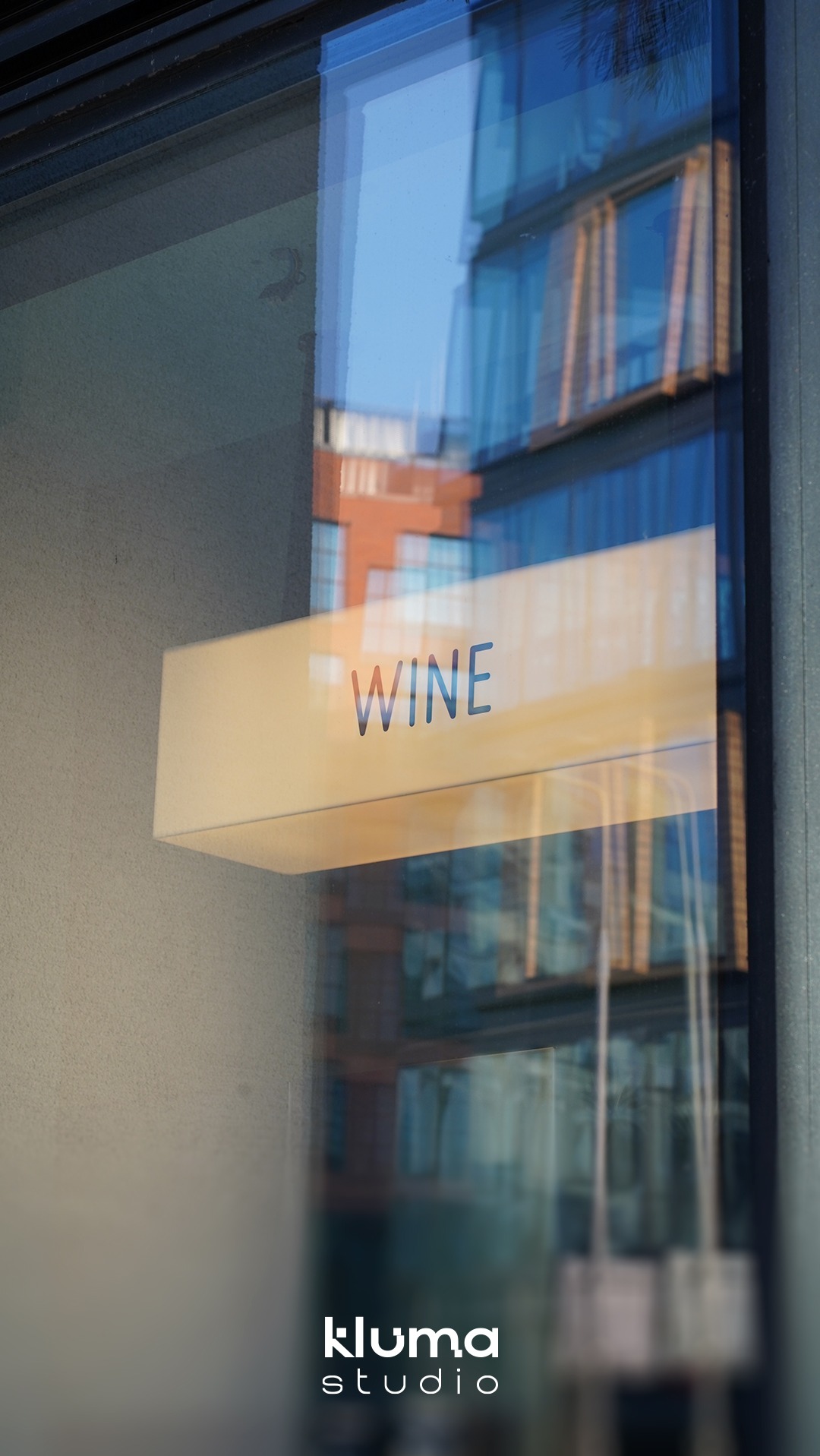 Podświetlany szyld z napisem 'WINE' zamontowany na szklanej fasadzie budynku, widoczny z perspektywy ulicy z odbiciami zabudowy miejskiej.